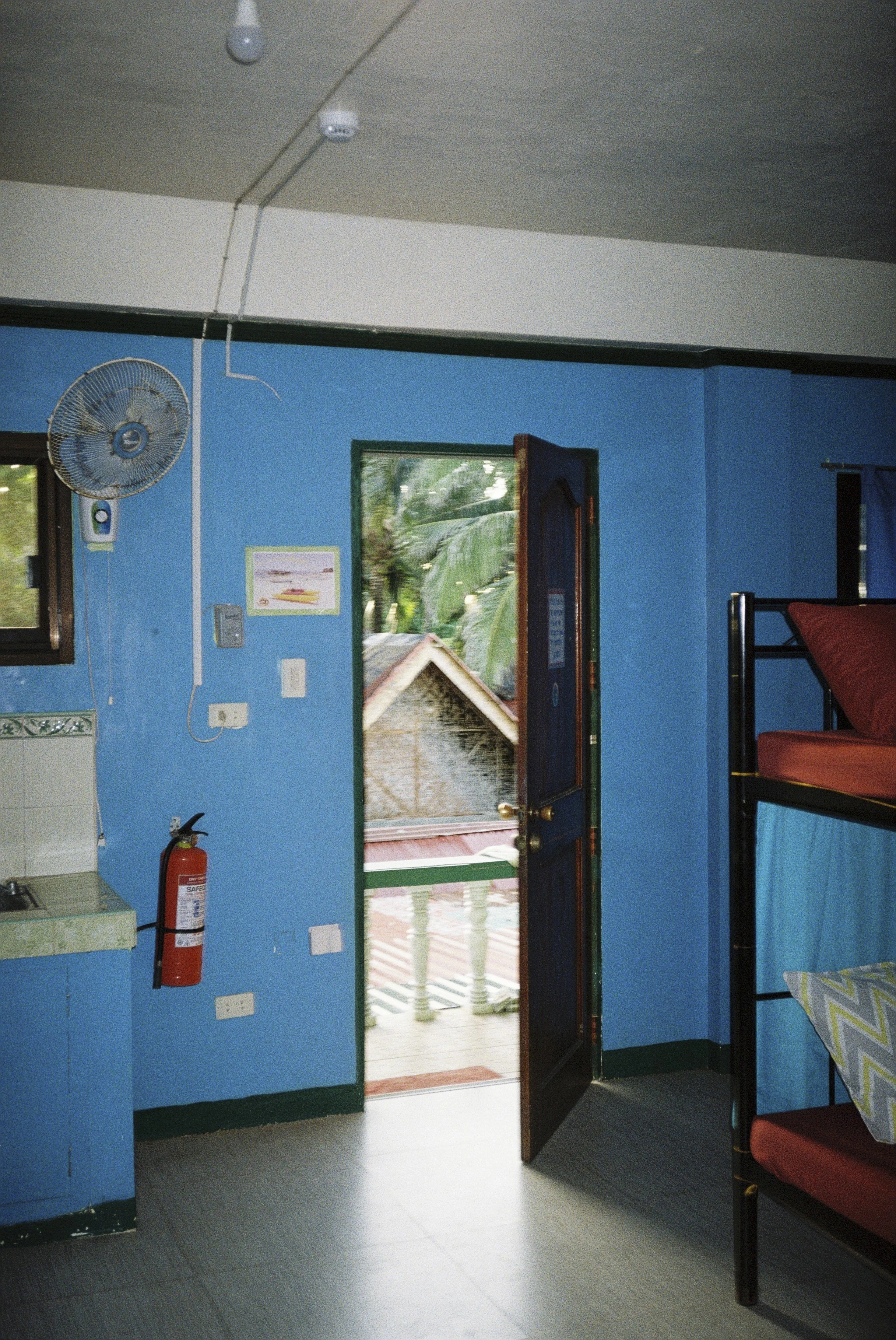 Blick in einen Raum mit blauer Wand, offener Tür, einem Fenster und einem roten Stuhl. Es gibt einen Feuerlöscher an der Wand, eine Deckenlampe und einen Wandventilator.