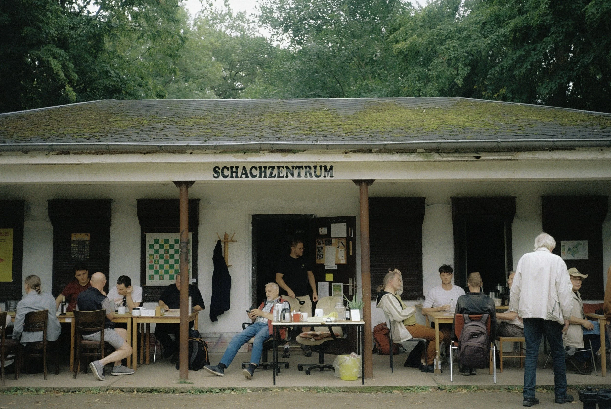 Menschen sitzen und stehen vor einem Gebäude mit dem Schild "Schachzentrum" auf dem Dach im Freien, umgeben von Bäumen.