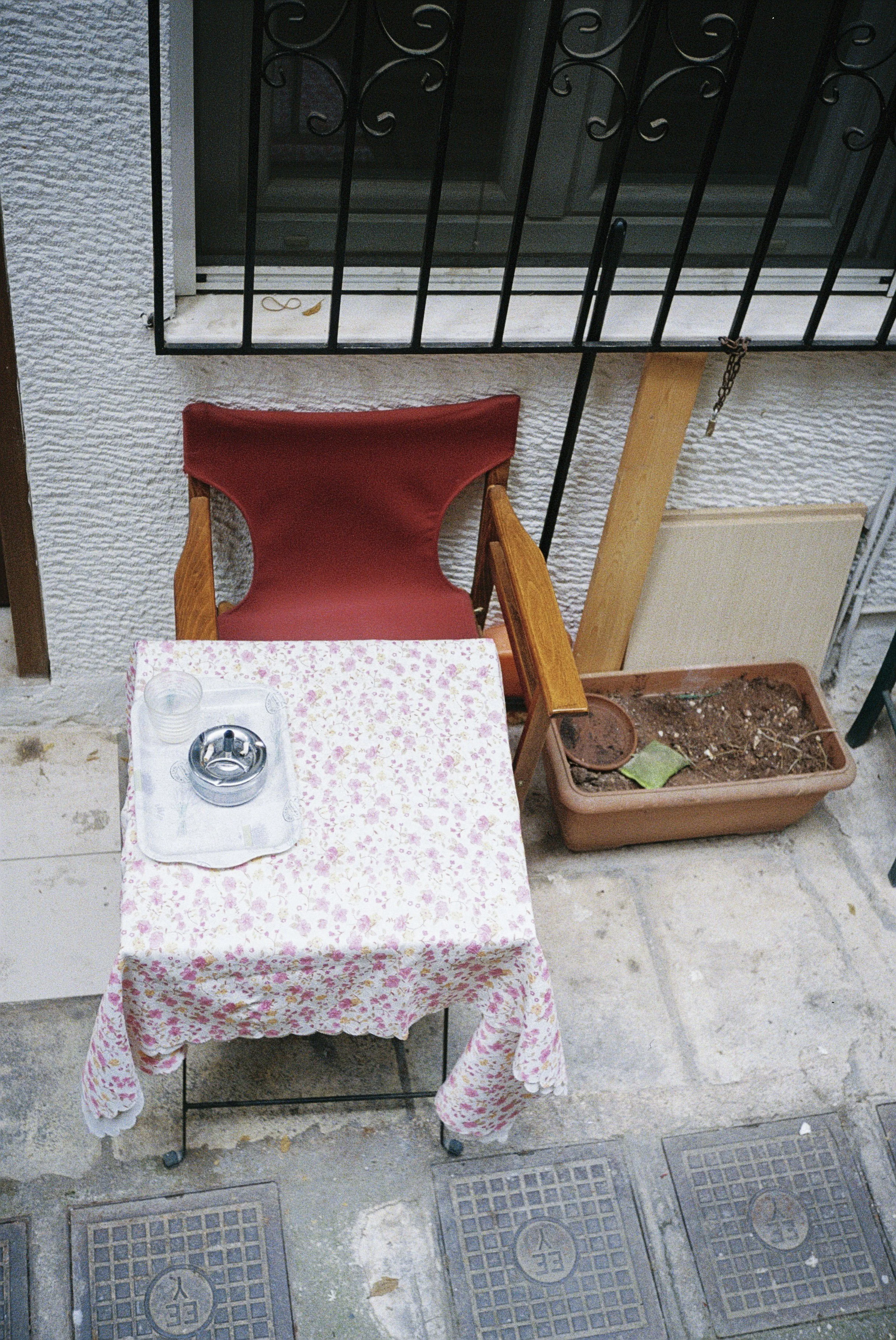 Ein Tisch mit einer weißen Tischdecke mit rosa Blumenmuster, darauf ein Aschenbecher, zwei leere Gläser, ein kleiner Salz- oder Pfefferstreuer, steht auf einem Bürgersteig neben einem Fenster mit schwarzen Metallgittern. Es gibt eine rote Stuhl und e
