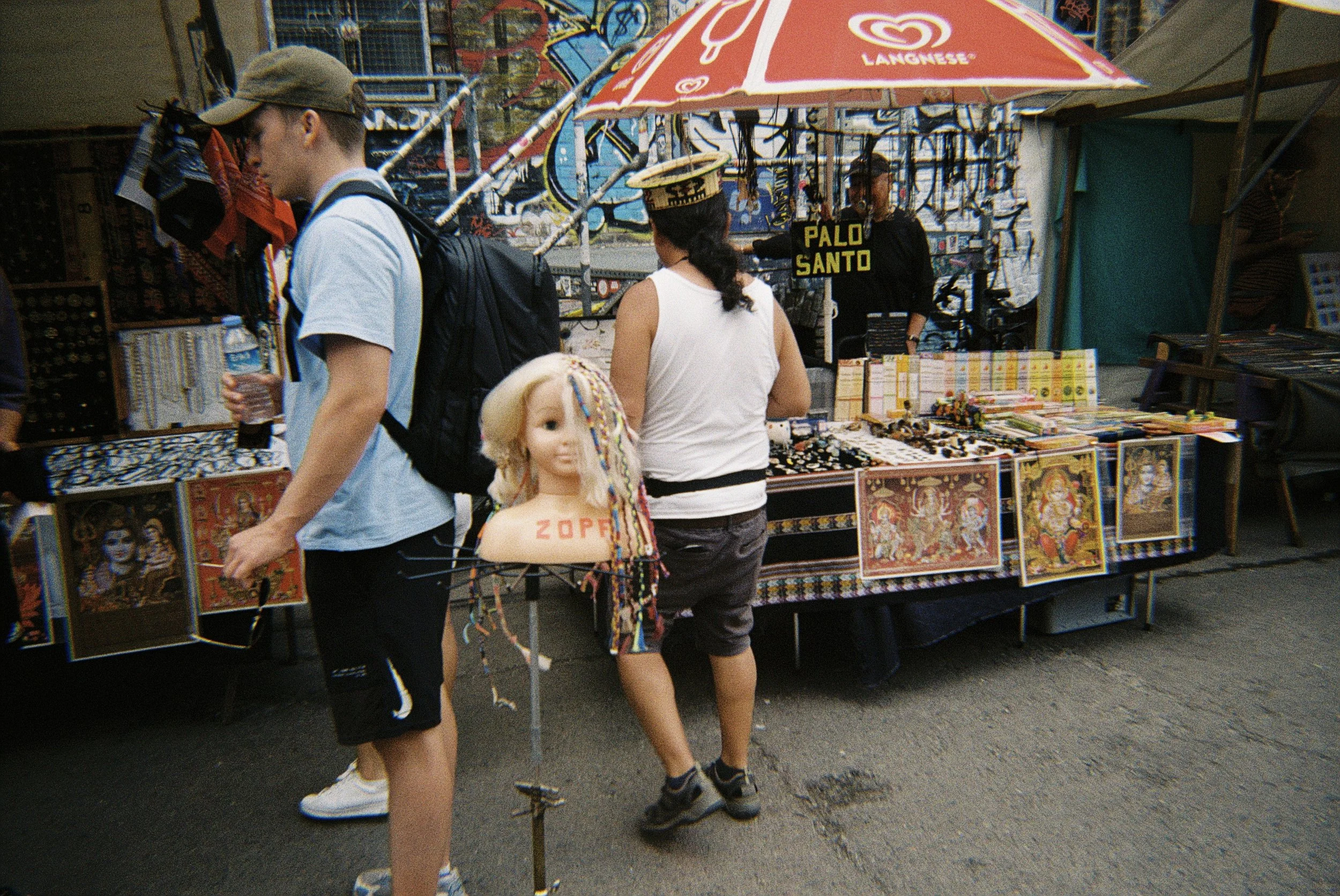 Menschen bei einem Straßenmarktstand, der Kunstwerke und Schmuck verkauft. Ein Unabhängiger mit einem Wasser in der Hand, ein Kind mit einem stingelbedeckten Blatt im Haar, und ein weiteres Mädchen mit einem schwarzen Kleid, das hinter dem Stand steh