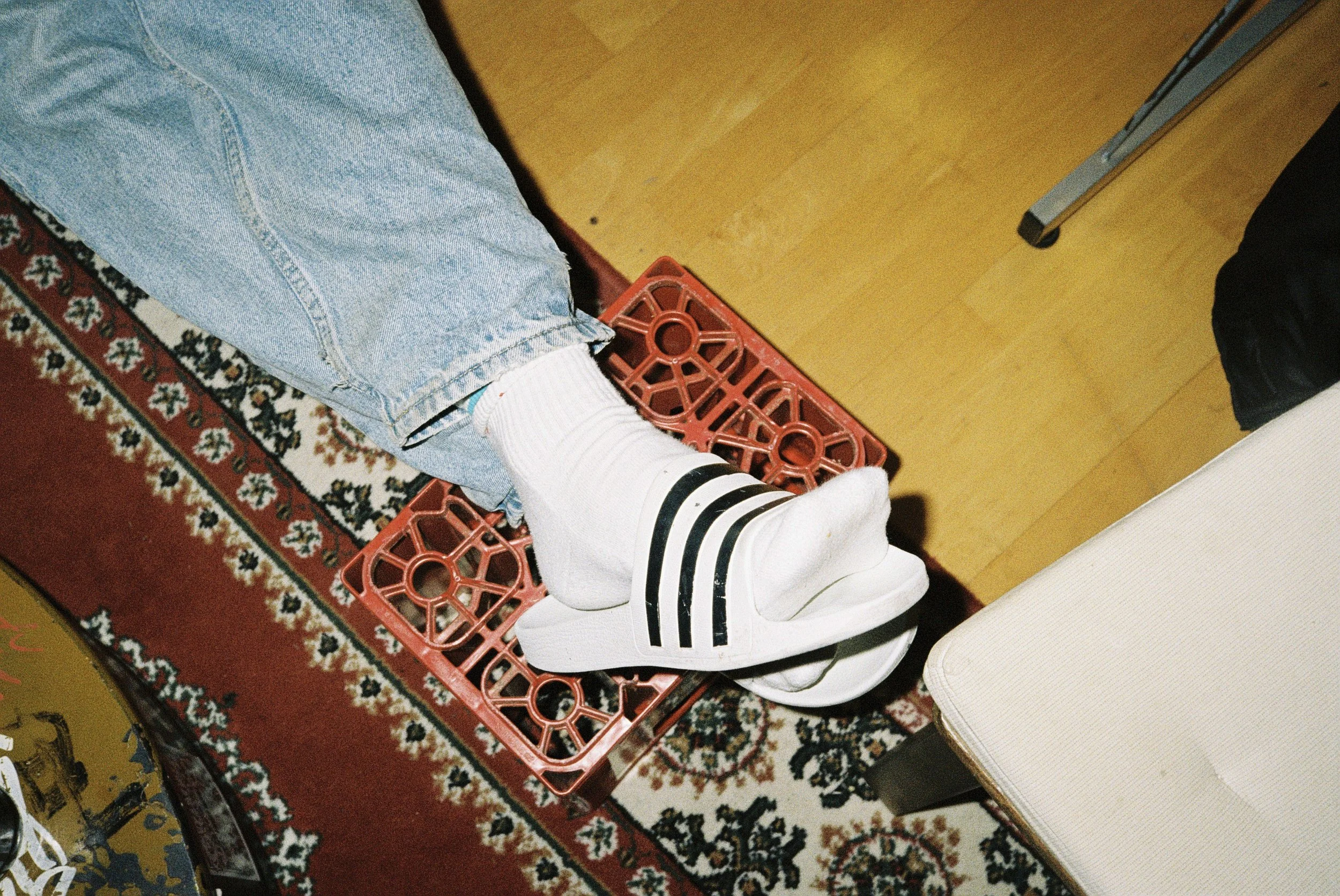 Person trägt weiße Sneakers mit schwarzen Streifen, sitzt auf einer roten Plastikablage, deren Beine auf einem Teppich mit Blumenmuster stehen, daneben Teil eines Schreibtisches und einer Holzfußboden.