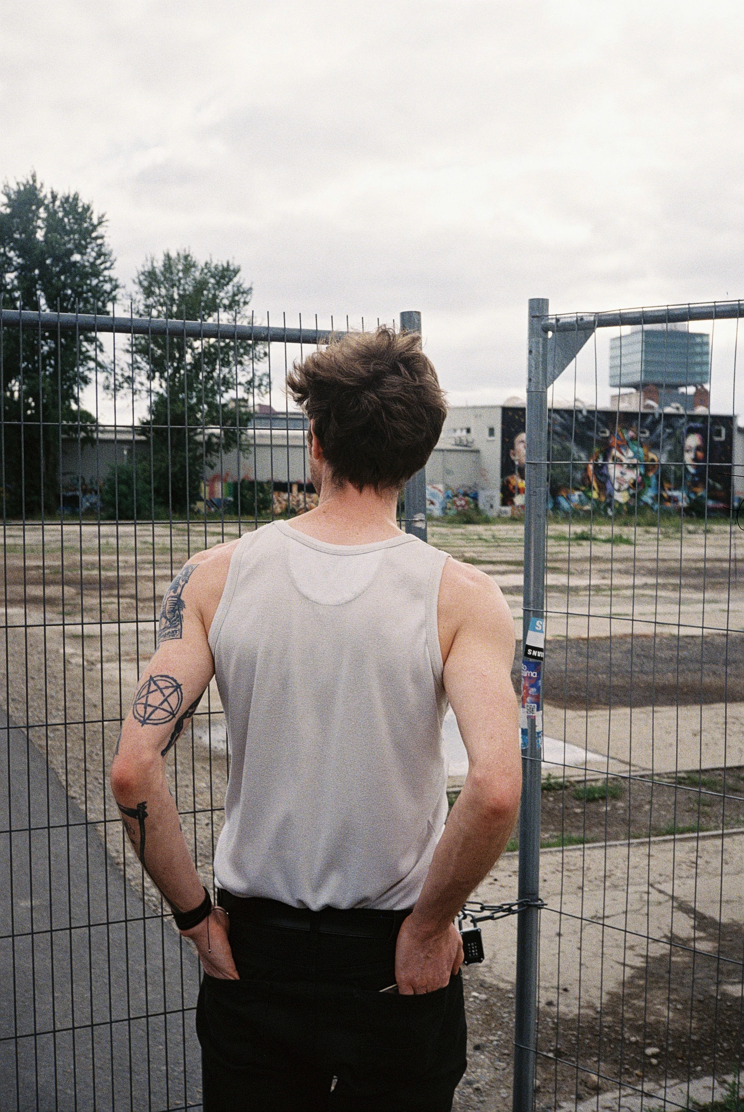 Der Rücken eines Mannes mit Tattoos, weißen Tanktop und schwarzen Hosen, der vor einem Drahtzaun steht, im Hintergrund Graffiti an einer Wand und Bäume bei bewölktem Himmel.