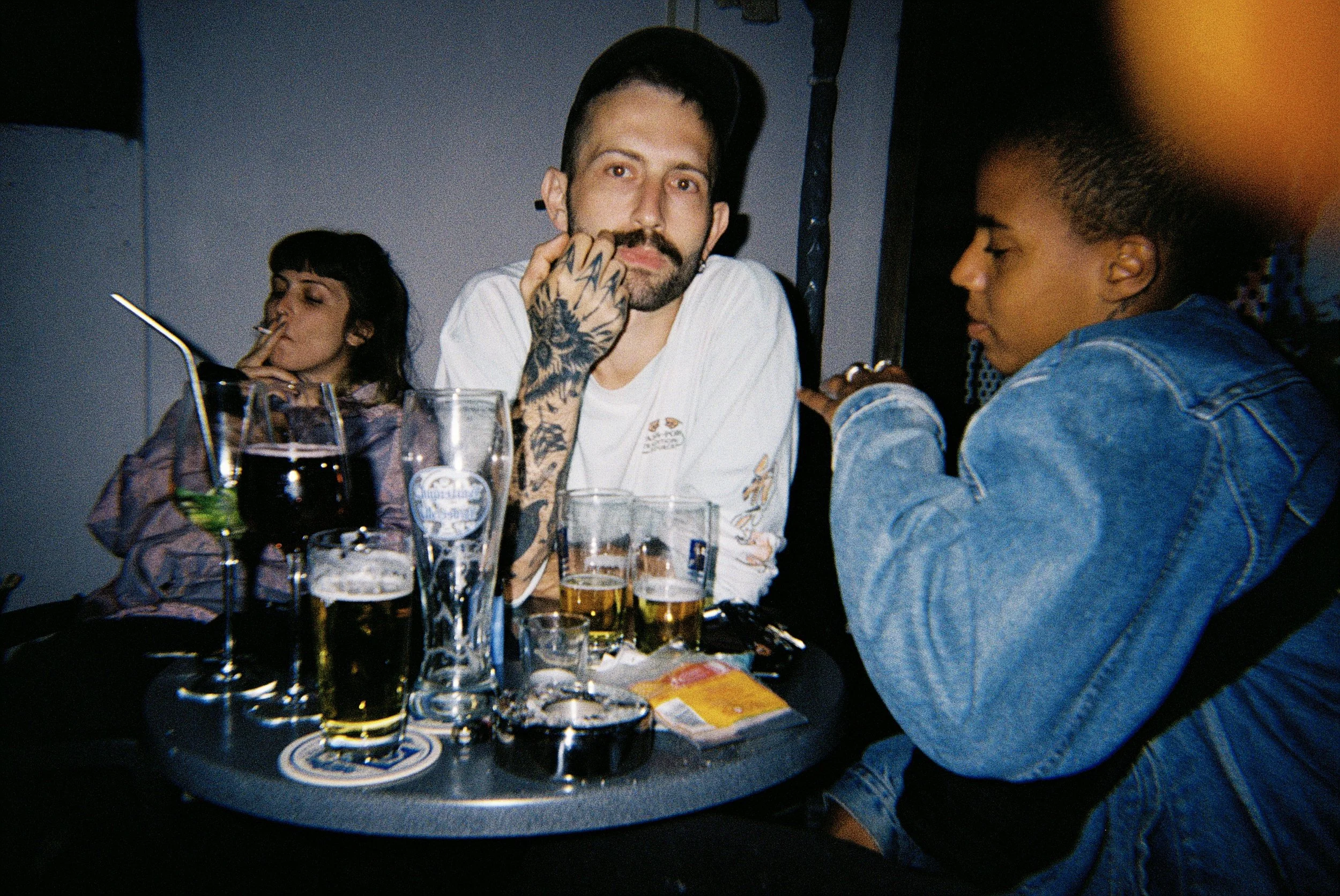 Menschen sitzen an einem Tisch mit Getränken und Snacks in einer Bar oder im Restaurant. Eine Person hat Tattoos am Arm und trägt ein Cap, während andere Personen neben ihr sitzen.