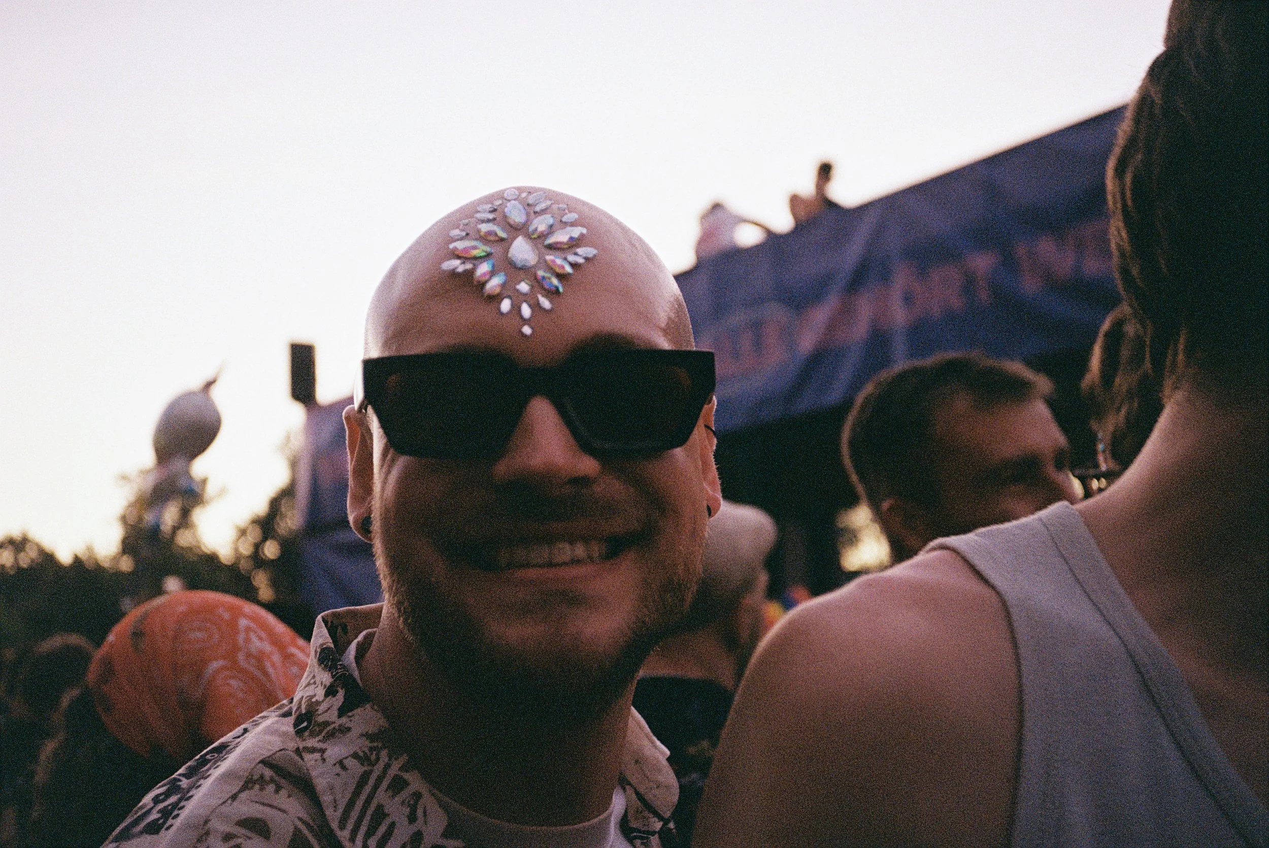 Ein lächelnder Mann mit Sonnenbrille und Glitzer-Kristallen auf der Stirn beim Festival, im Hintergrund andere Menschen und ein Zeltdach.
