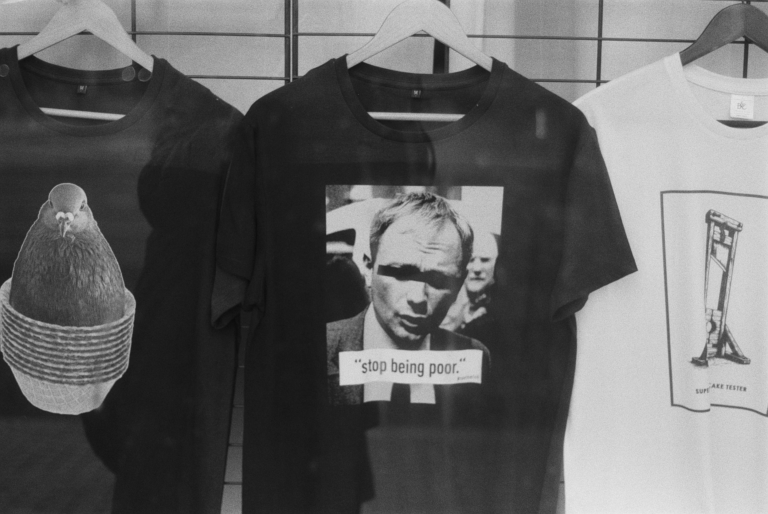 Schwarz-weiß Foto von T-Shirts auf Kleiderbügel, eines mit einem Portrait und einem Zitat, eines mit einem Prominentengesicht, ein weiteres mit einer Hand und einem Werkzeug, und ein T-Shirt mit einem Schild, das 'stop being poor' sagt. Es gibt auch 
