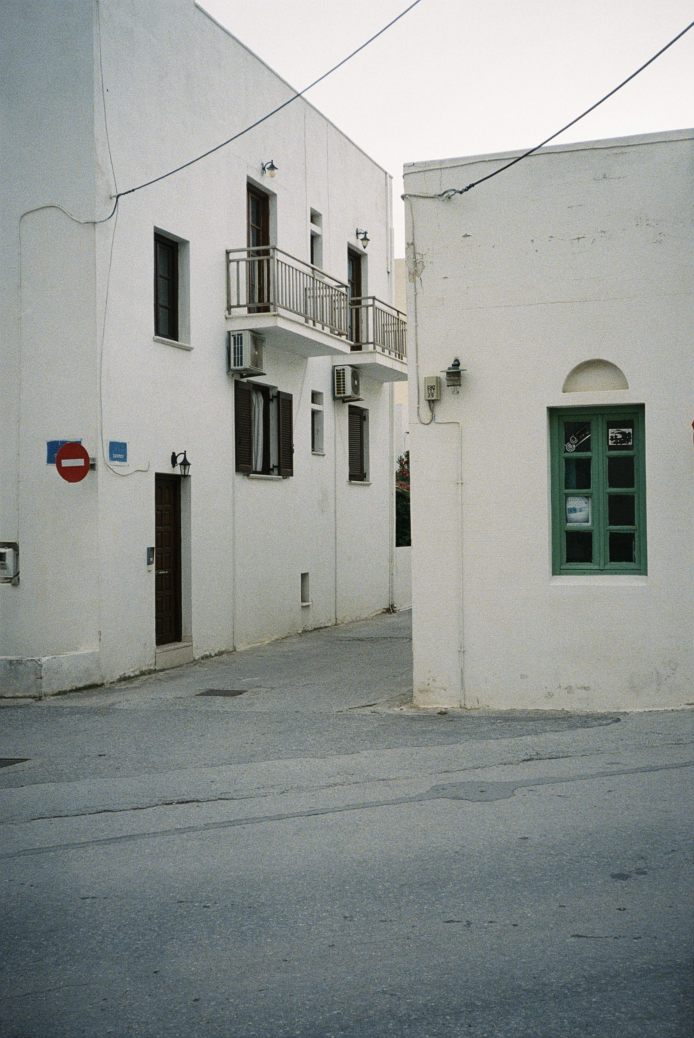 Weiße Gebäude mit dunklen Fensterläden und Balkonen, grüne Tür, Stromkabel, graue Straße in einer ruhigen, scheinbar mediterranen Gegend.