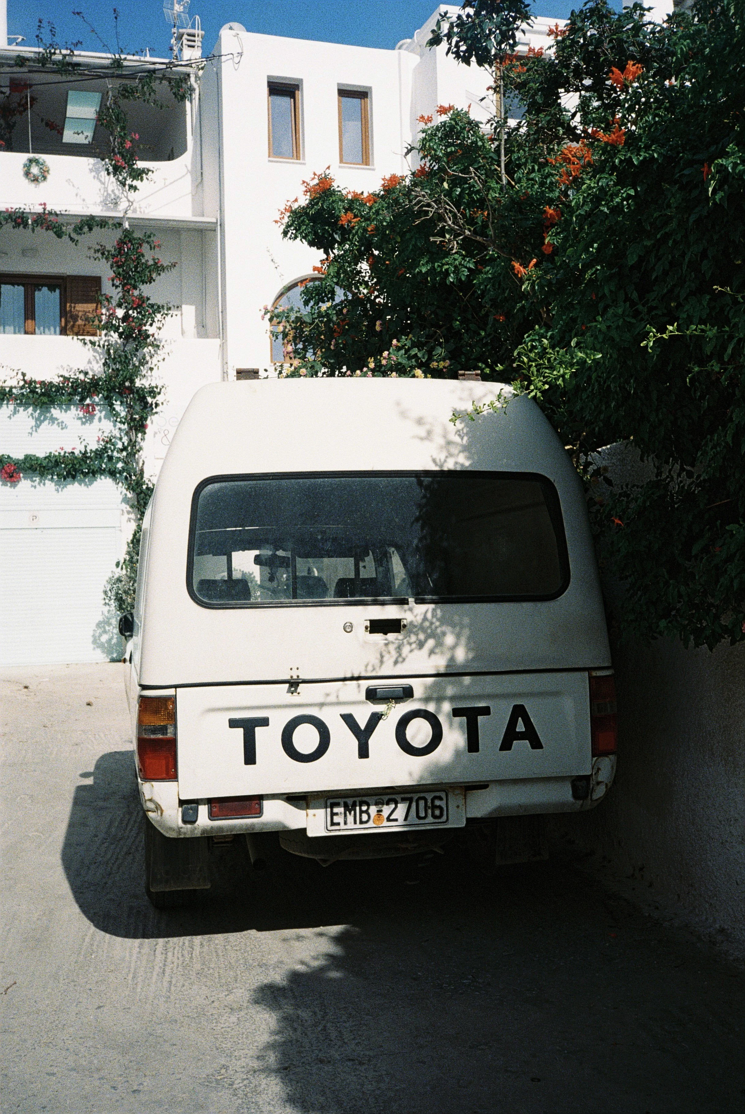 Weiße Toyota-Transporter mit deutschem Kennzeichen, vor einer weißen Hausfassade mit Fenstern, im Hintergrund ein Baum mit orange Blüten, auf der Straße geparkt.