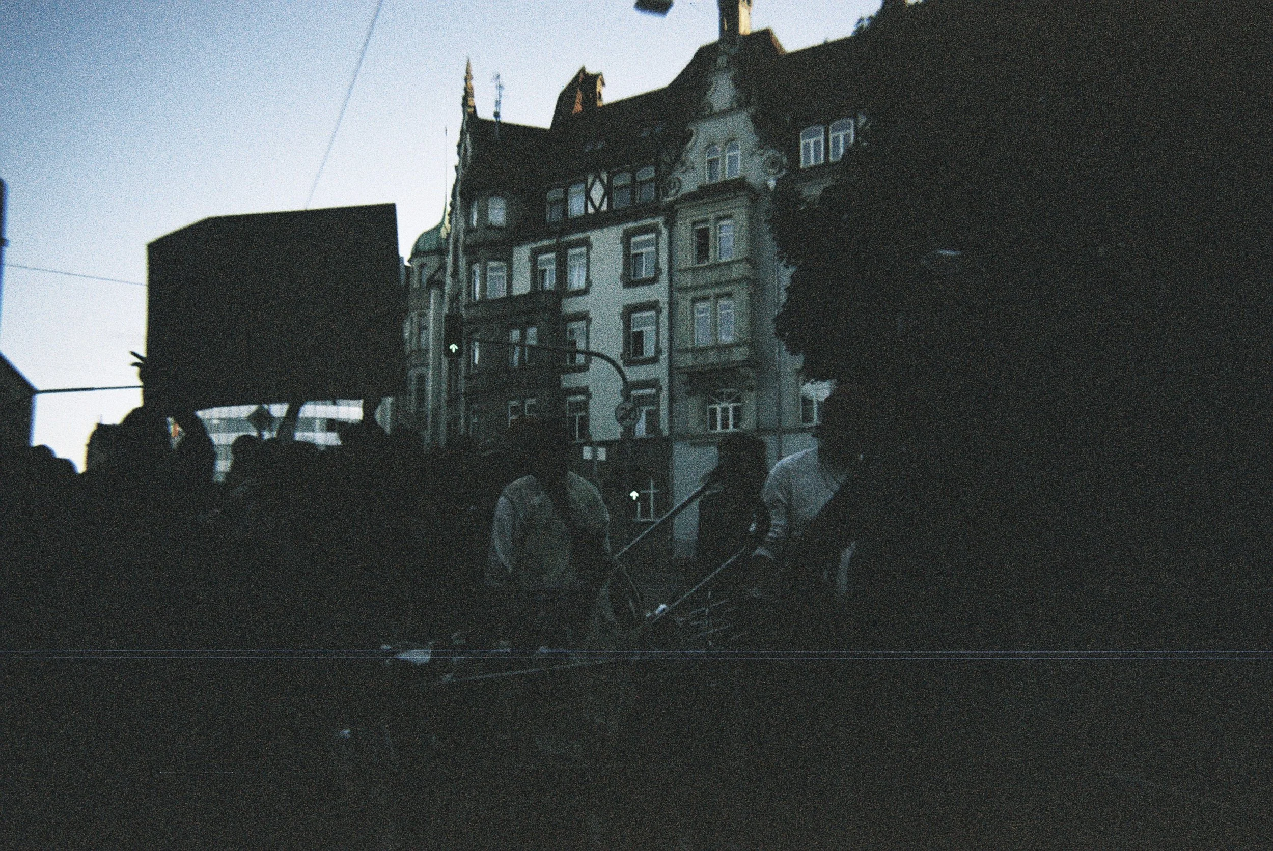 Dunkle, unscharfe Stadtaufnahme mit mehreren Menschen im Vordergrund und historischen Gebäuden im Hintergrund bei Dämmerung.