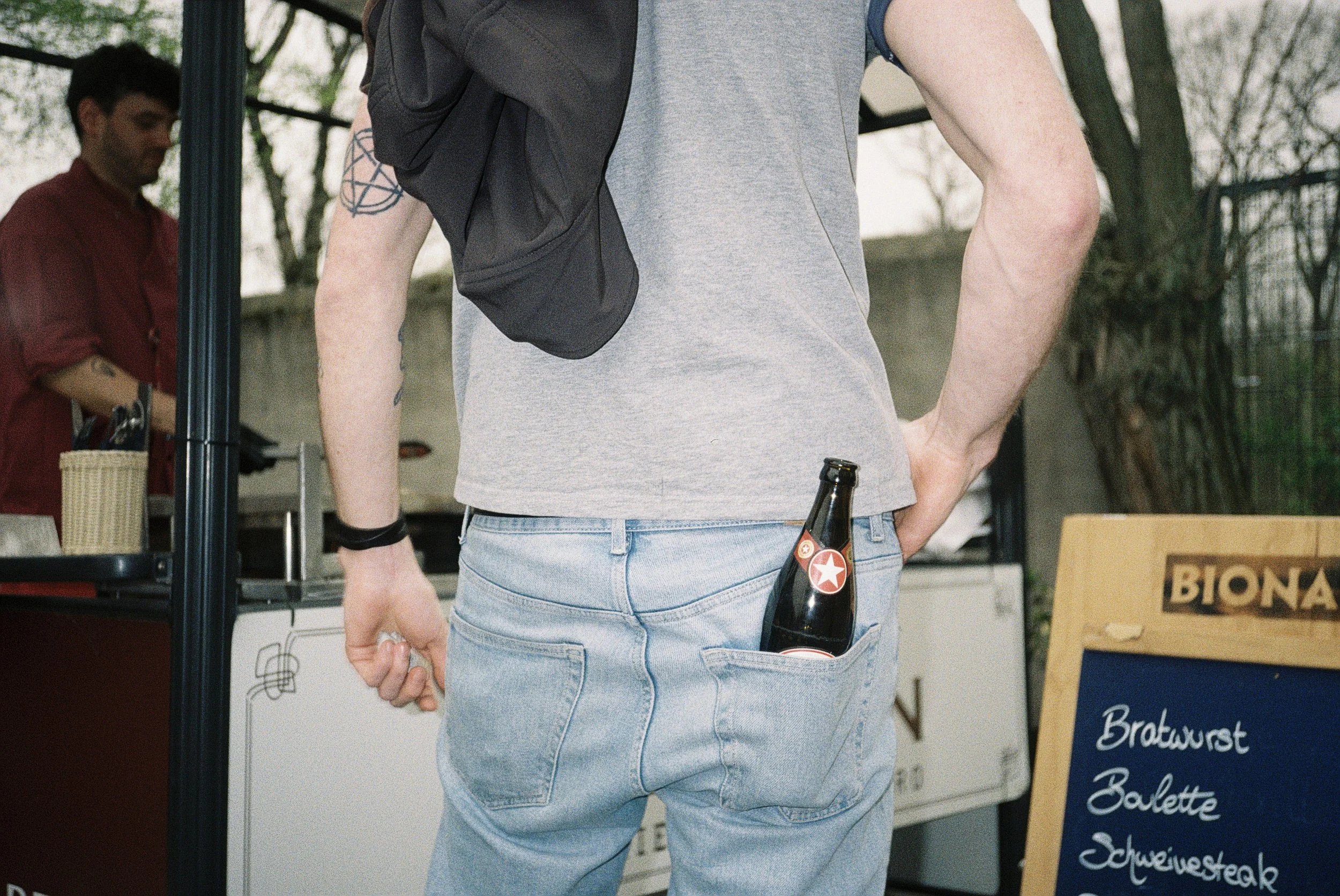 Person in grauem T-Shirt mit einer schwarzen Tasche auf der Schulter, die Hand in die Tasche stecken, in einem Markt mit einem Getränkeflasche in der Hosentasche und einem Mann im roten Hemd im Hintergrund.