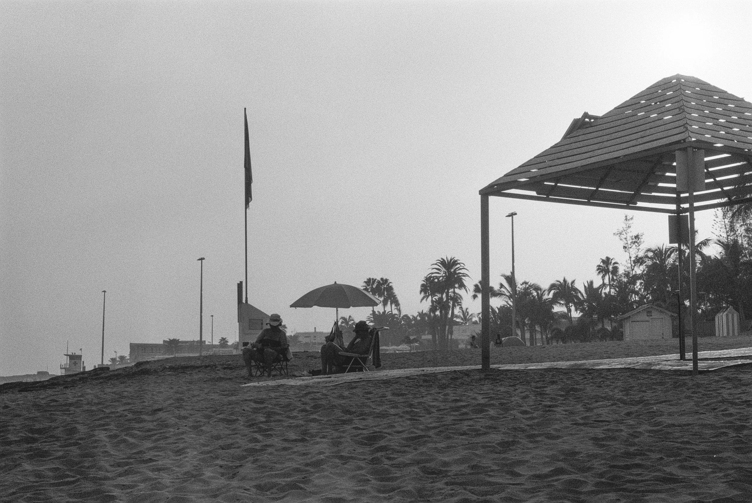 Schwarz-weiß-Foto vom Strand mit Palmen, Menschen im Schatten unter Sonnenschirmen und einer Holzpergola.