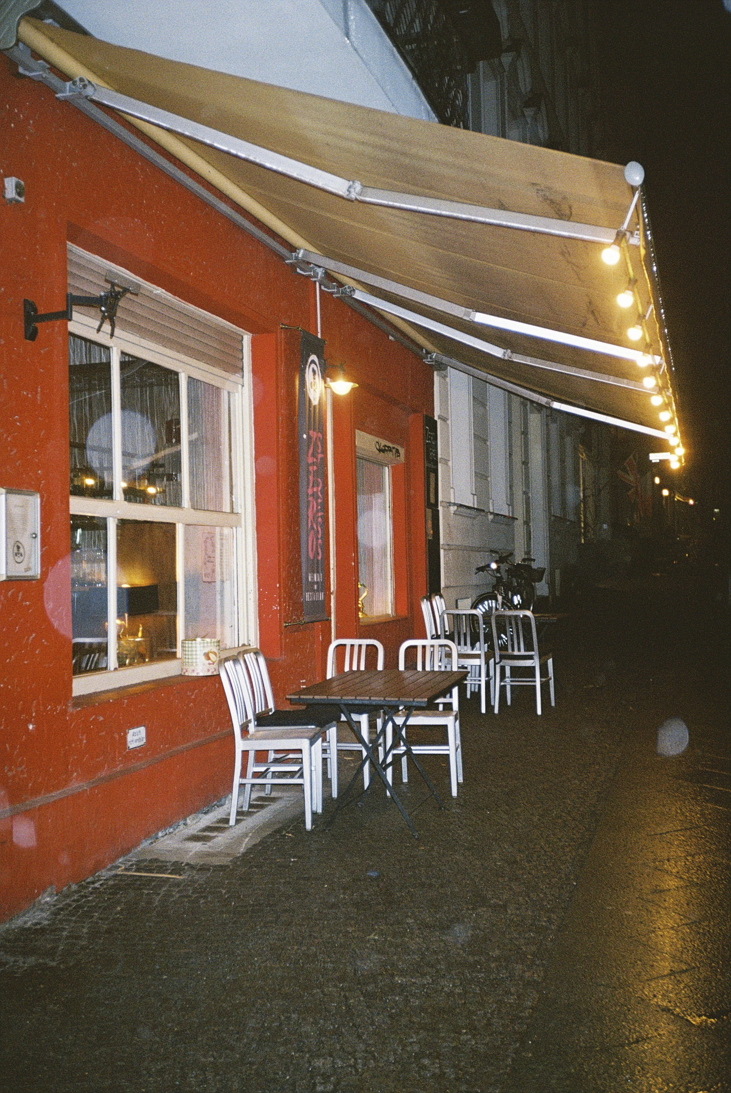 Außenaufnahme eines Restaurants oder Cafés bei Nacht mit Tischen und Stühlen auf dem Gehweg, roter Fassadenwand, Fenstern und Beleuchtung. Es sieht ruhig und einladend aus.