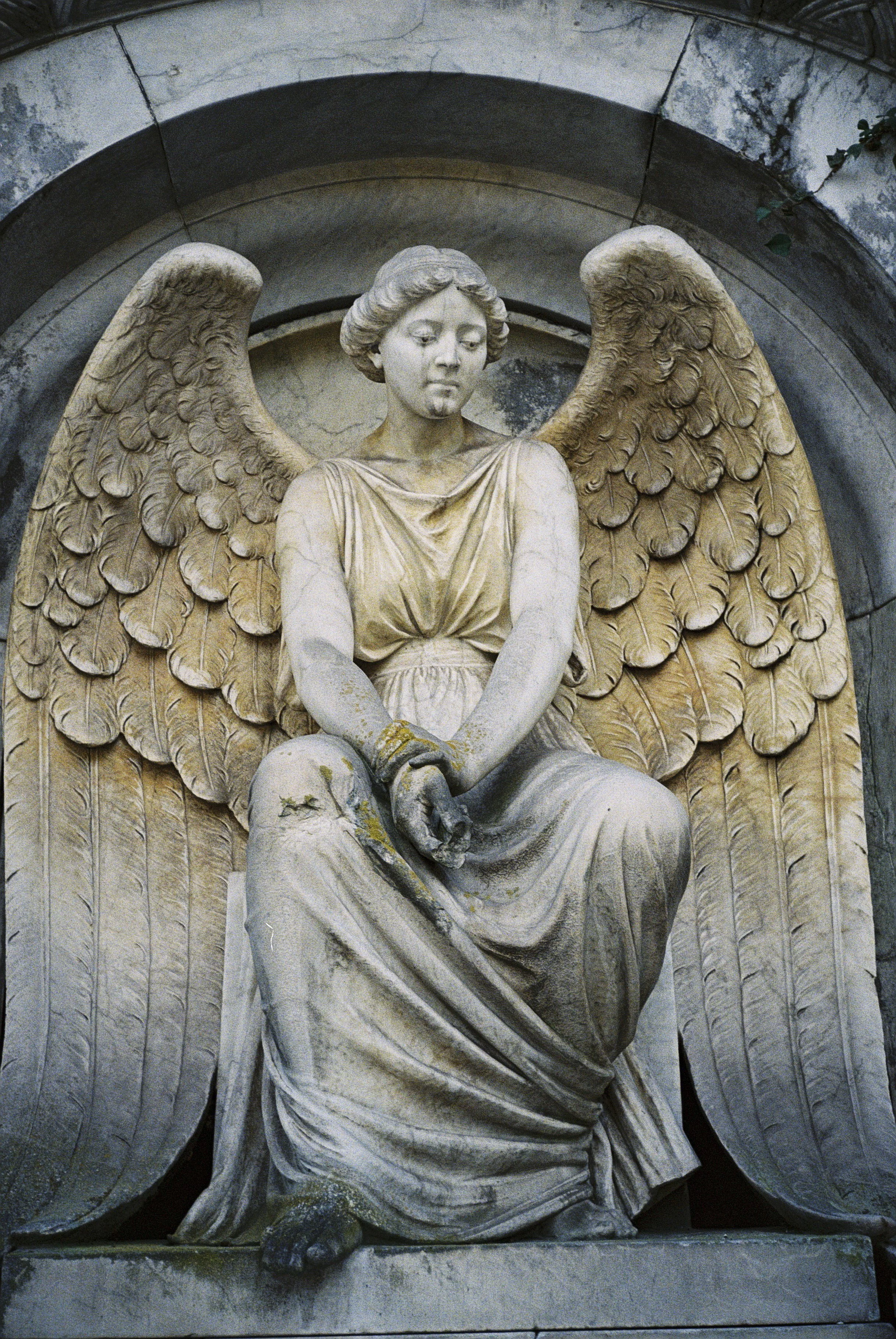 Steinerne Skulptur eines Engels mit großen Flügeln, der sitzt, die Hände auf den Oberschenkeln, mit einer nachdenklichen Miene.
