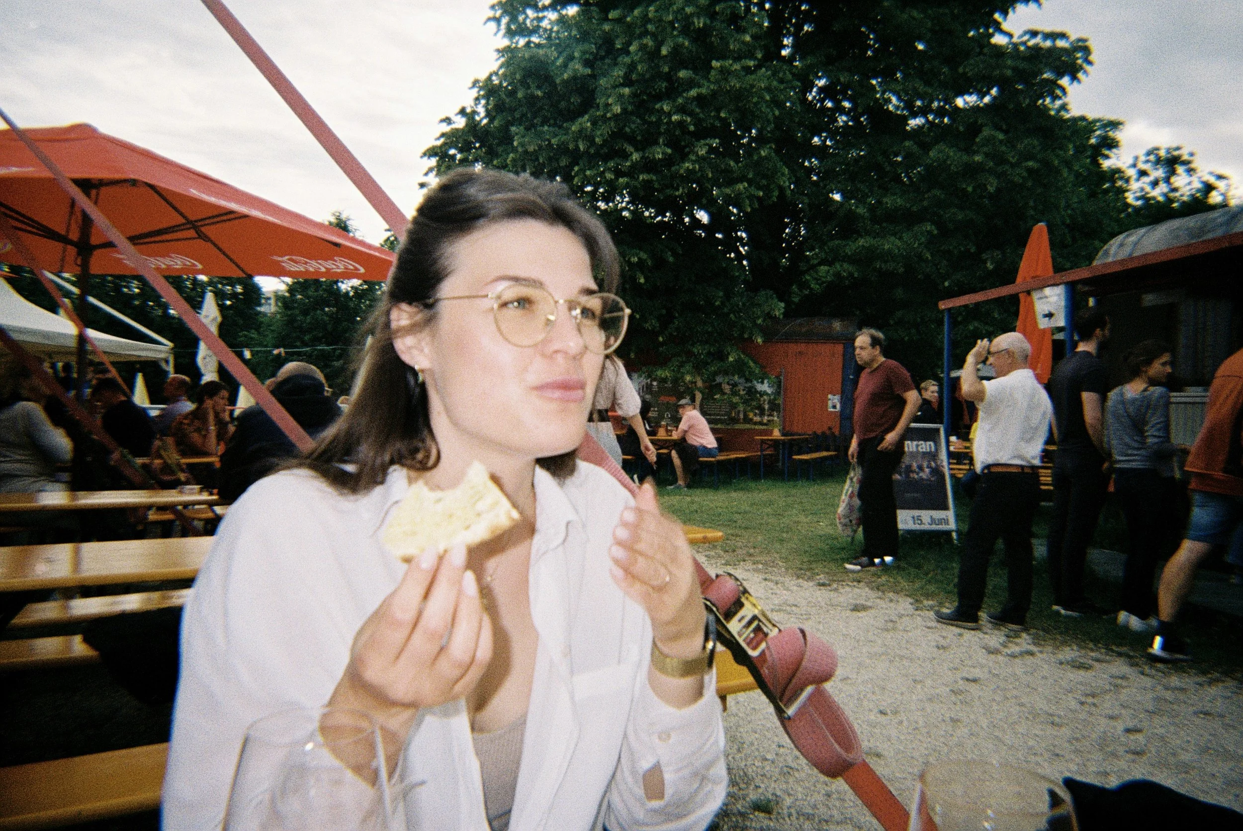 Eine junge Frau mit Brille und weißen Hemd isst ein Stück Kuchen oder Flan auf einer äußeren Sitzgelegenheit. Im Hintergrund sind mehrere Leute, Bänke, Schirme und ein Baum, was auf eine öffentliche Veranstaltung oder ein Fest im Freien hinweist.