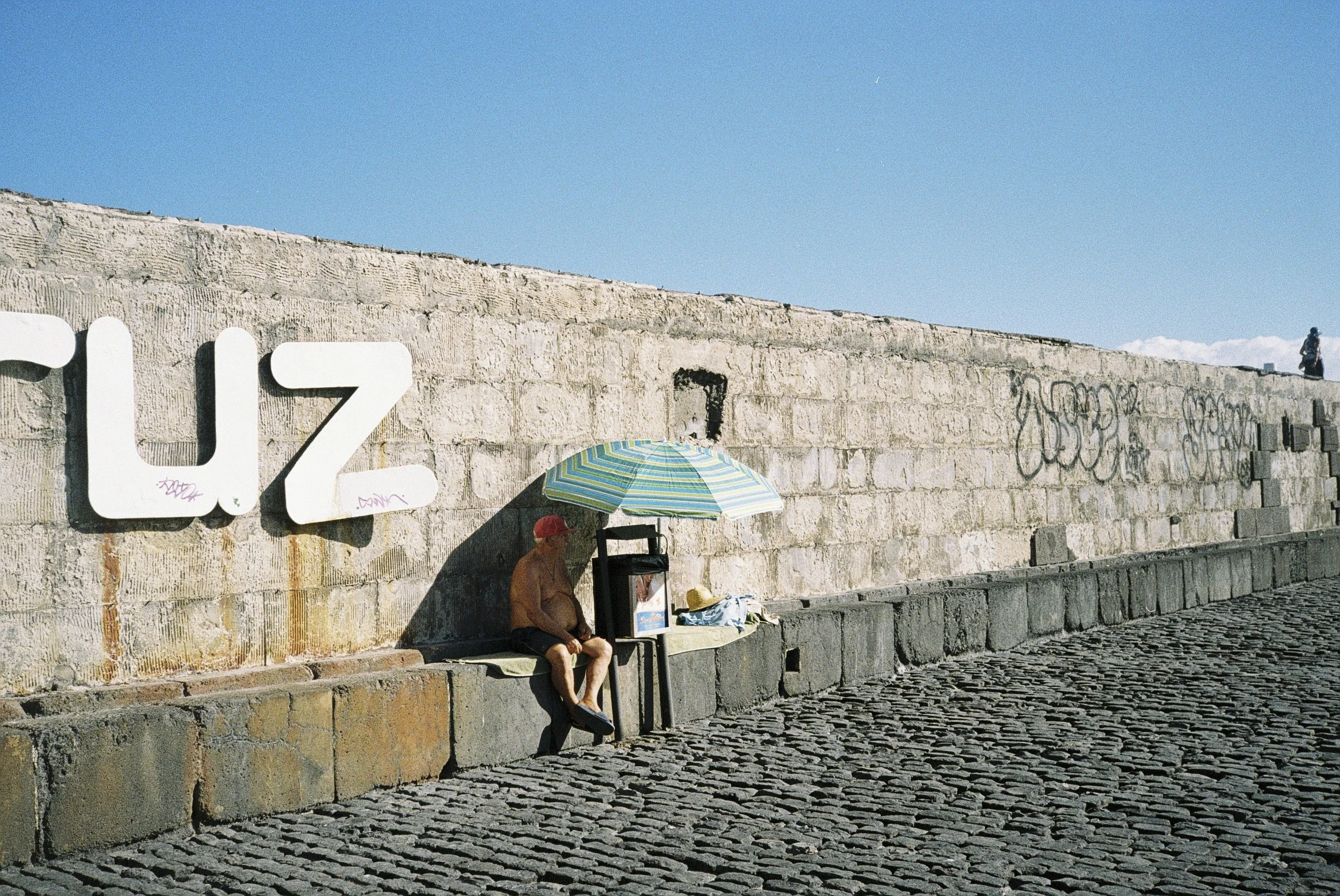Ein Mann sitzt am Flussufer auf einer Bank, neben ihm liegen Sachen, im Hintergrund eine hohe Steinmauer mit Graffiti, ein Mensch mit einem Regenschirm steht vor der Mauer, der Himmel ist blau mit wenigen Wolken.