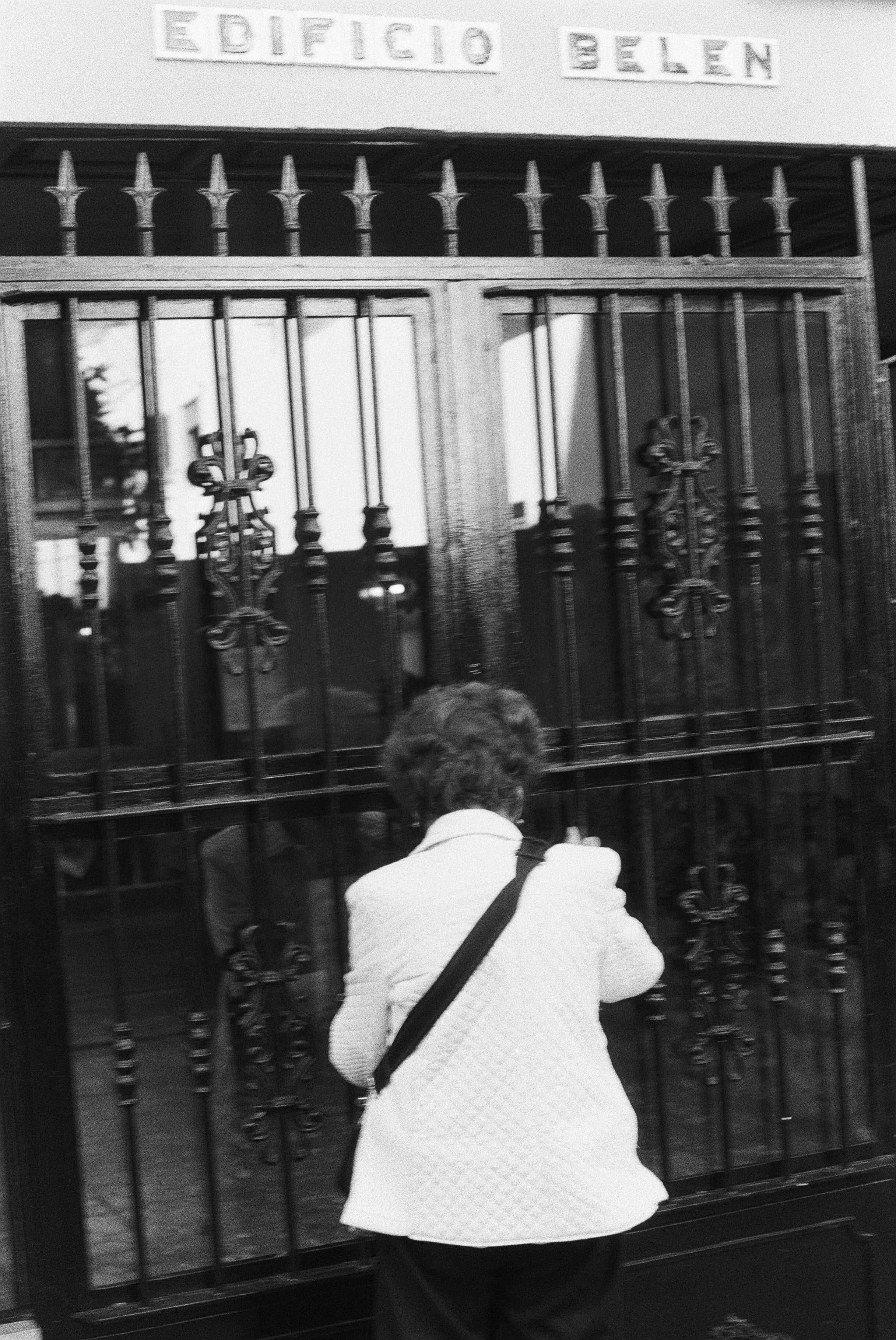 Eine Person mit weißen Jacke und schwarzer Tasche vor einem metallischen Tor, das mit Griffen und Dekorationen verziert ist. Am oberen Bereich ist ein Schild mit der Aufschrift 'EDIFICIO Belen' zu sehen.