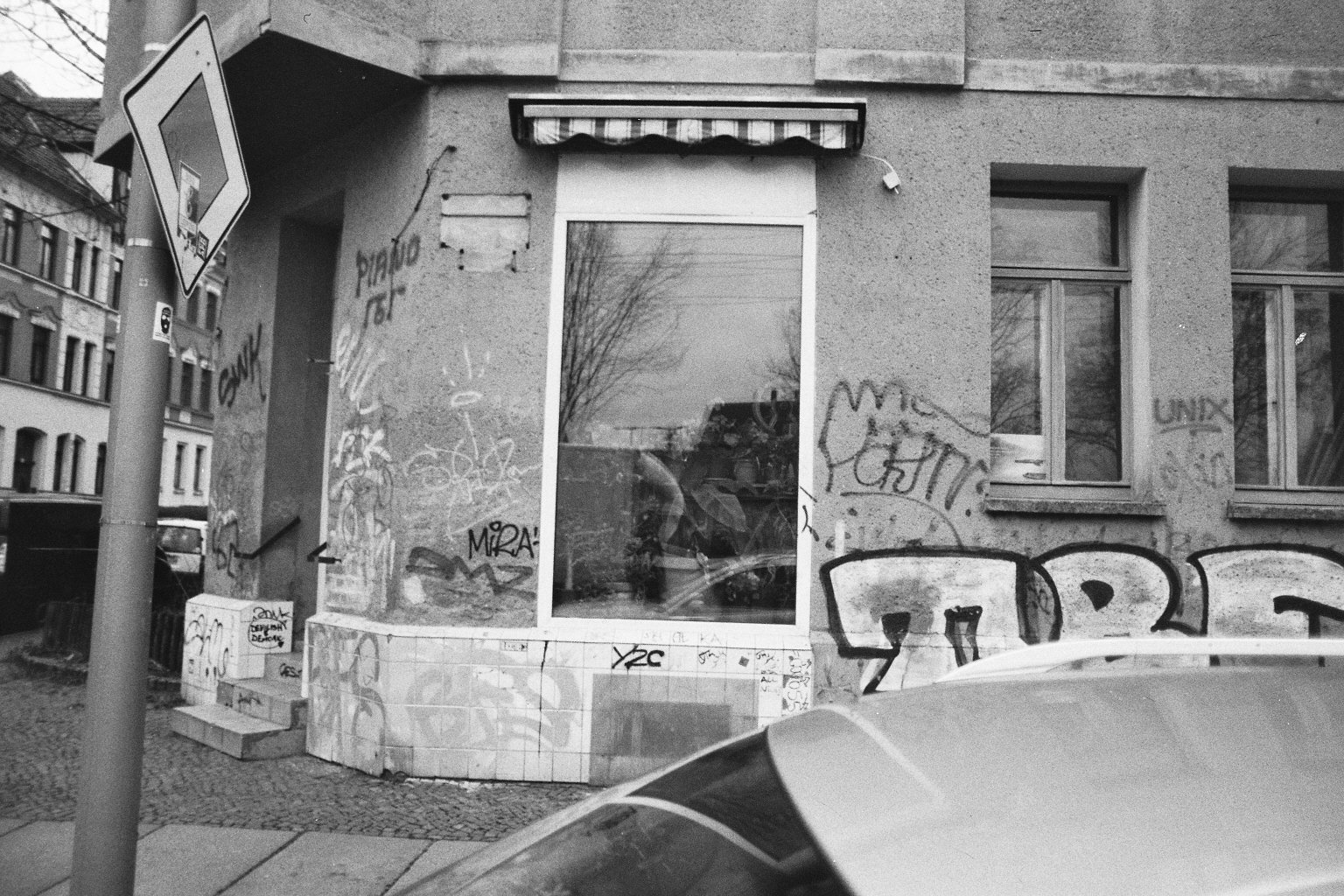 Ein graues Gebäude mit Graffiti an der Wand, mehreren Fenstern und einem Eingang. Im Vordergrund ist ein Teil eines geparkten Autos sichtbar, außerdem ein Parkverbotsschild.
