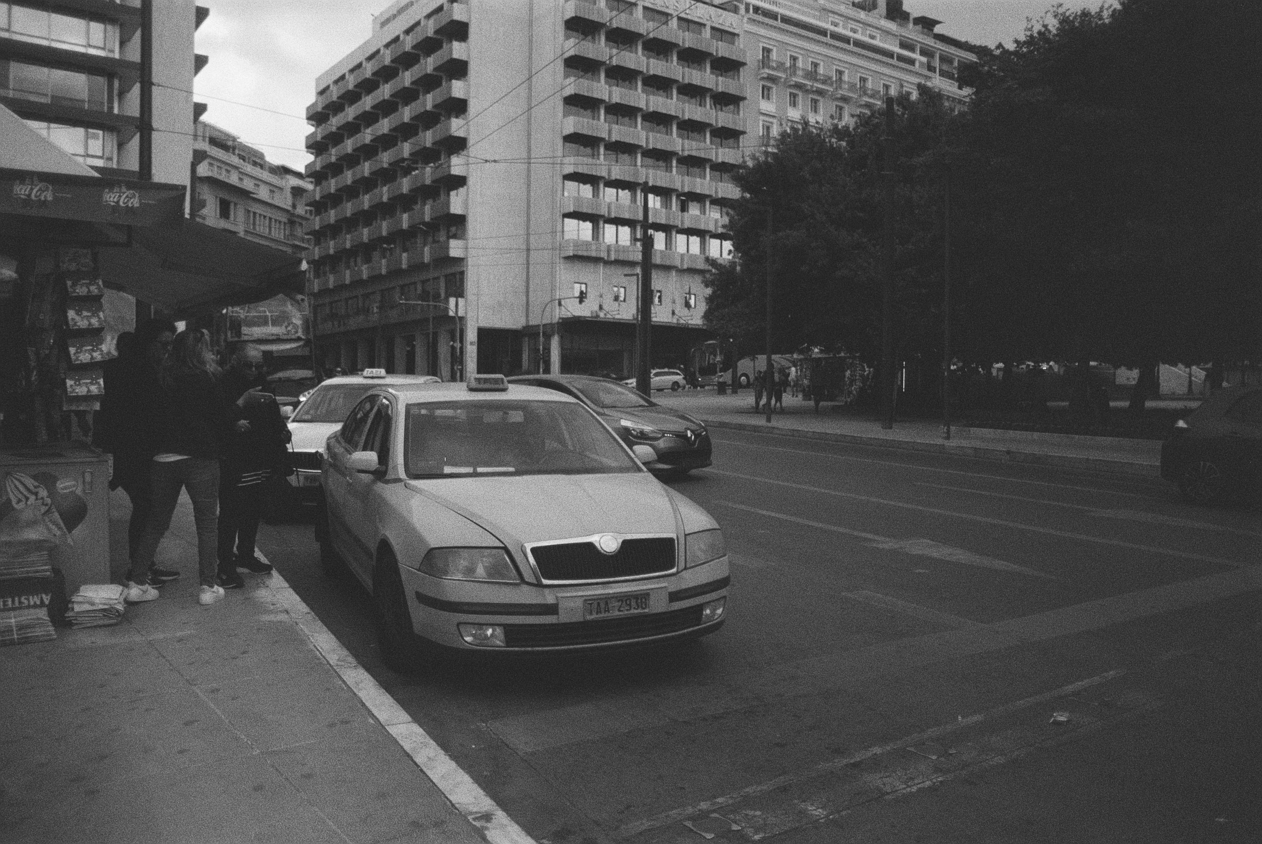 Straßenansicht mit mehreren Autos, Hochhäuser im Hintergrund und Menschen auf dem Bürgersteig.