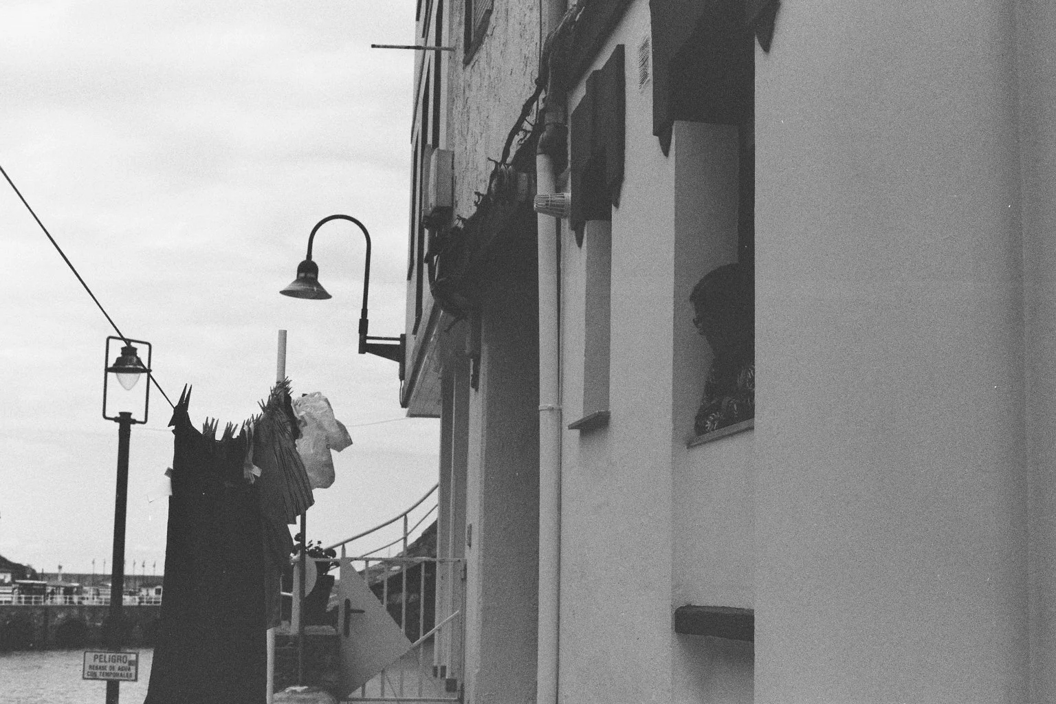 Schwarz-Weiß-Foto eines Hauses mit Waschlein auf dem Balkon im Vordergrund, Klamotten hängen zum Trocknen auf einer Leine, Straßenlaternen und Wasser im Hintergrund.