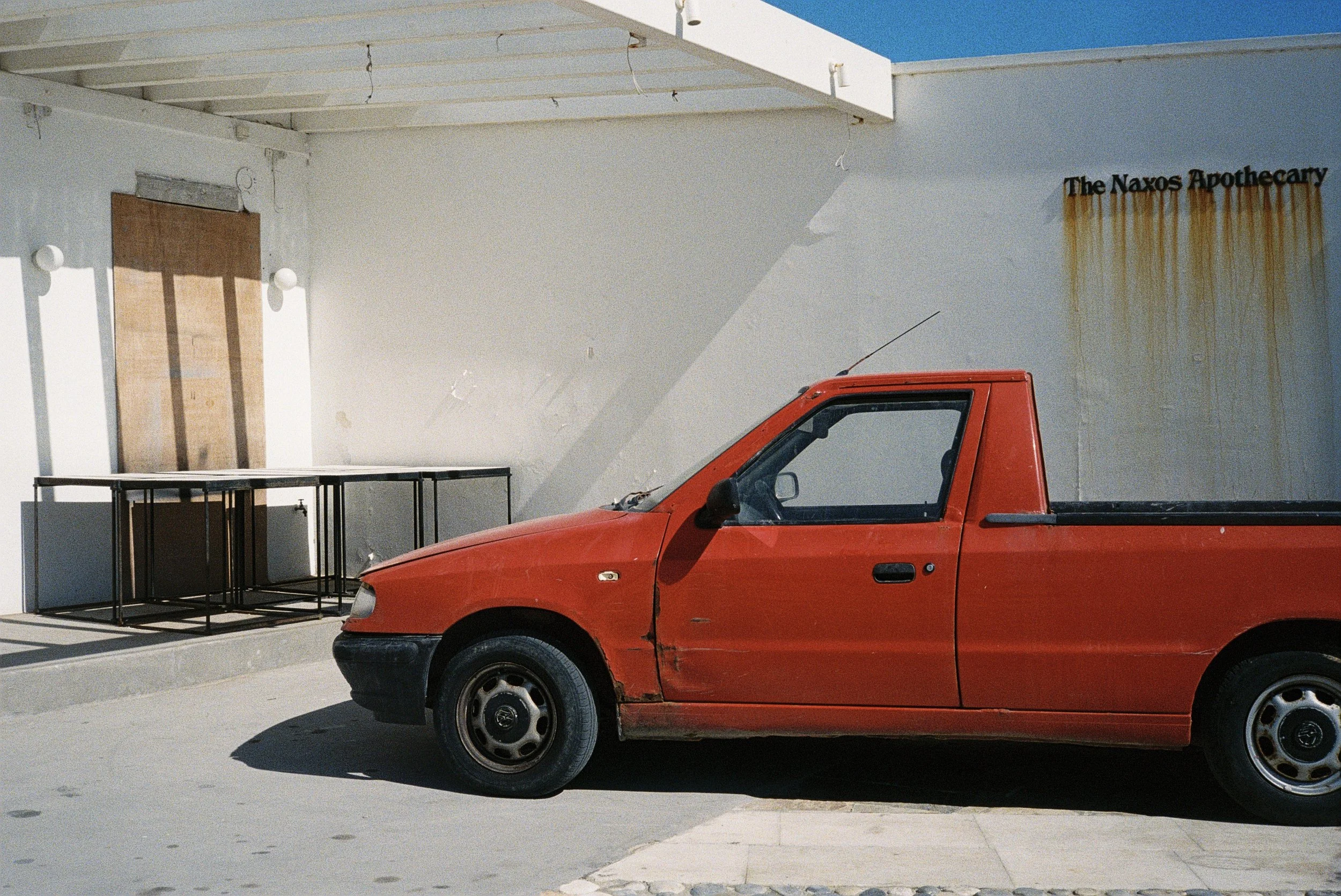 Ein rotes Auto, das vor einer weißen Wand steht, mit einer Holzverkleidung am oberen Rand und einem rustikalen Rustfleck. Es gibt mehrere schwarze Tische und eine Tür, die kaum sichtbar ist.