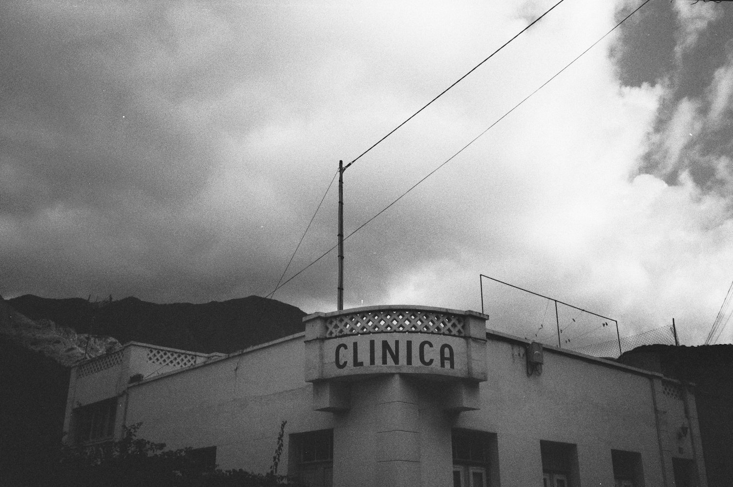 Schwarzes und weißes Foto einer Klinik mit dem Schriftzug 'CLINICA' auf einem Gebäude. Im Hintergrund Berge und Wolken am Himmel.