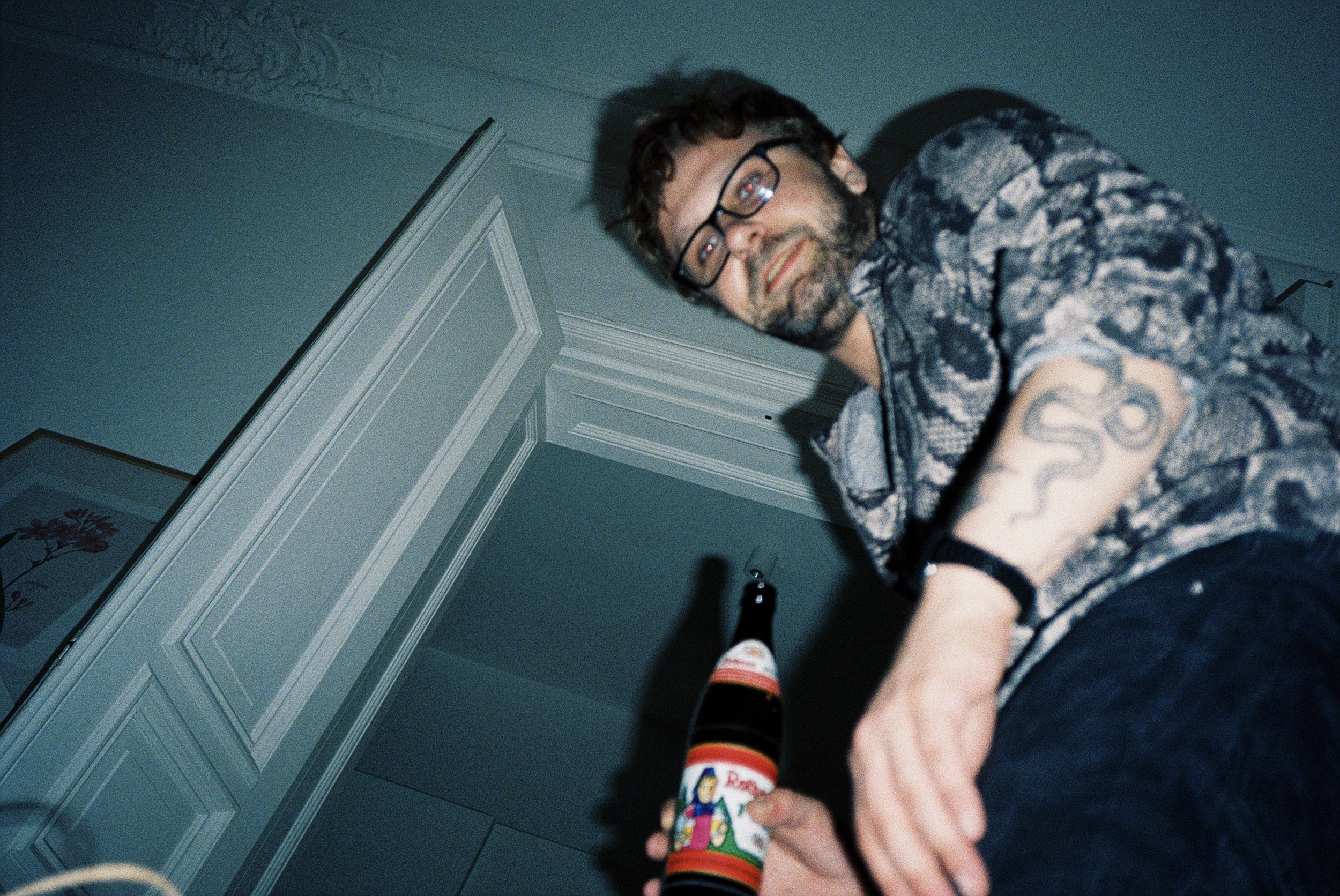 Ein Mann mit Brille und Bart hält eine Flasche mit weihnachtlichem Etikett in der Hand, während er in einem Raum mit weißen Wänden steht.