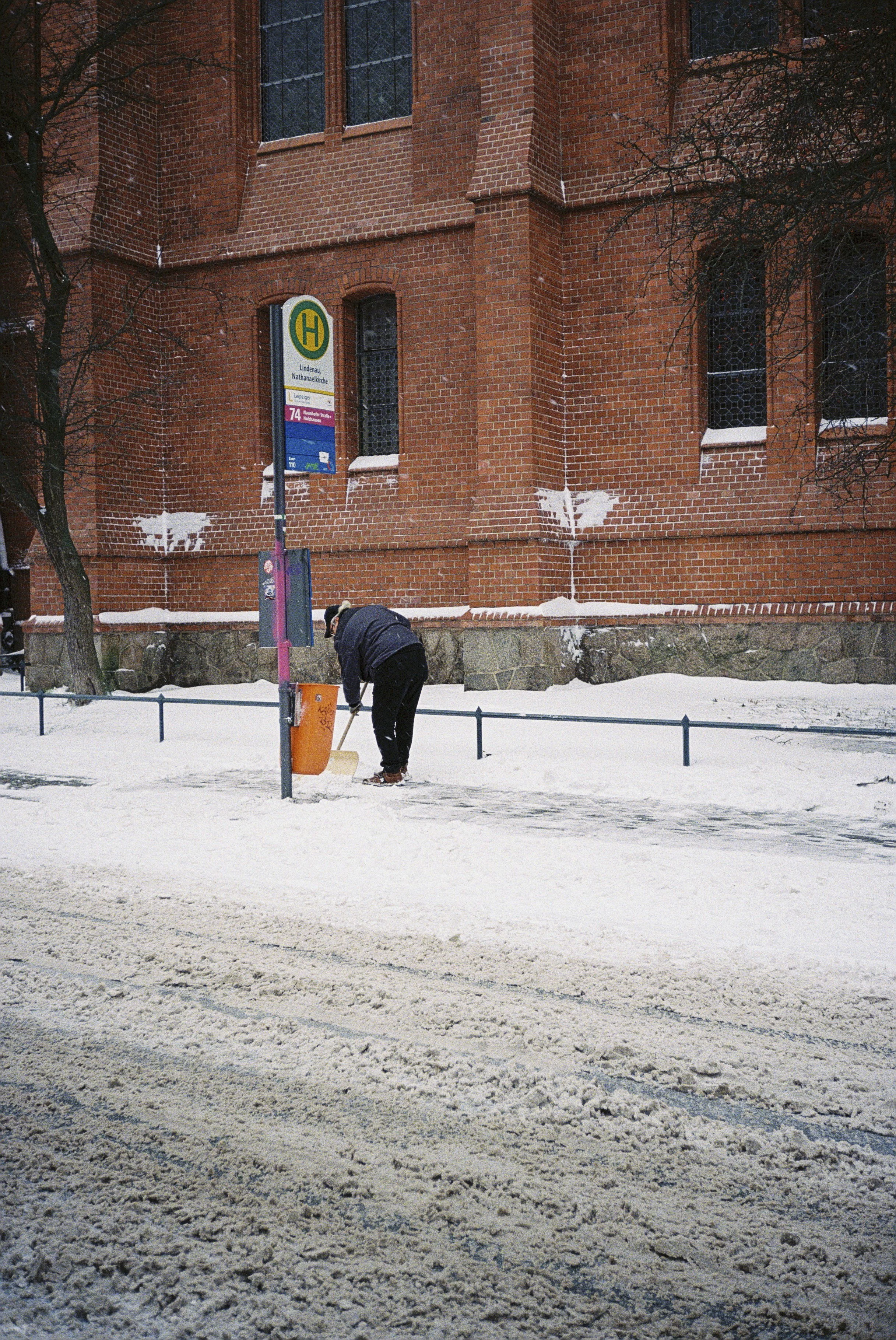 Eine Person kehrt Schnee auf einem Gehweg vor einem roten Backsteingebäude, neben einem Bushaltestellen-Schild, im Winter.