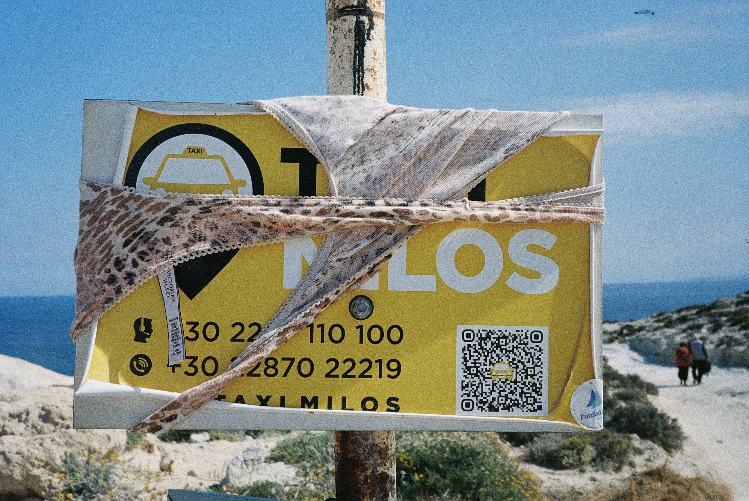 Gelbe Taxi-Preistafel am Strand, teilweise bedeckt mit einem Baby- und einem Tuch, mit QR-Code und Kontaktinformationen, am Strand bei sonnigem Wetter.