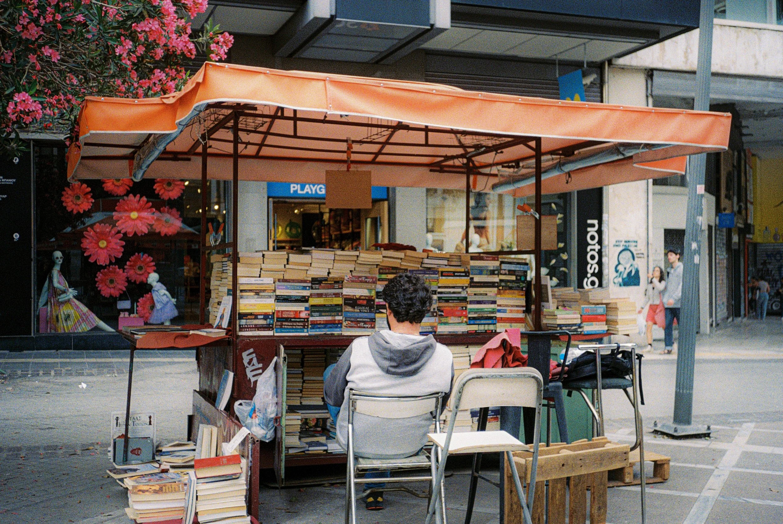 Straßenverkaufsstand mit Büchern vor einem Geschäft, eingerahmt von pinken Blumen, im urbanen Gebiet, mit mehreren Menschen im Hintergrund.