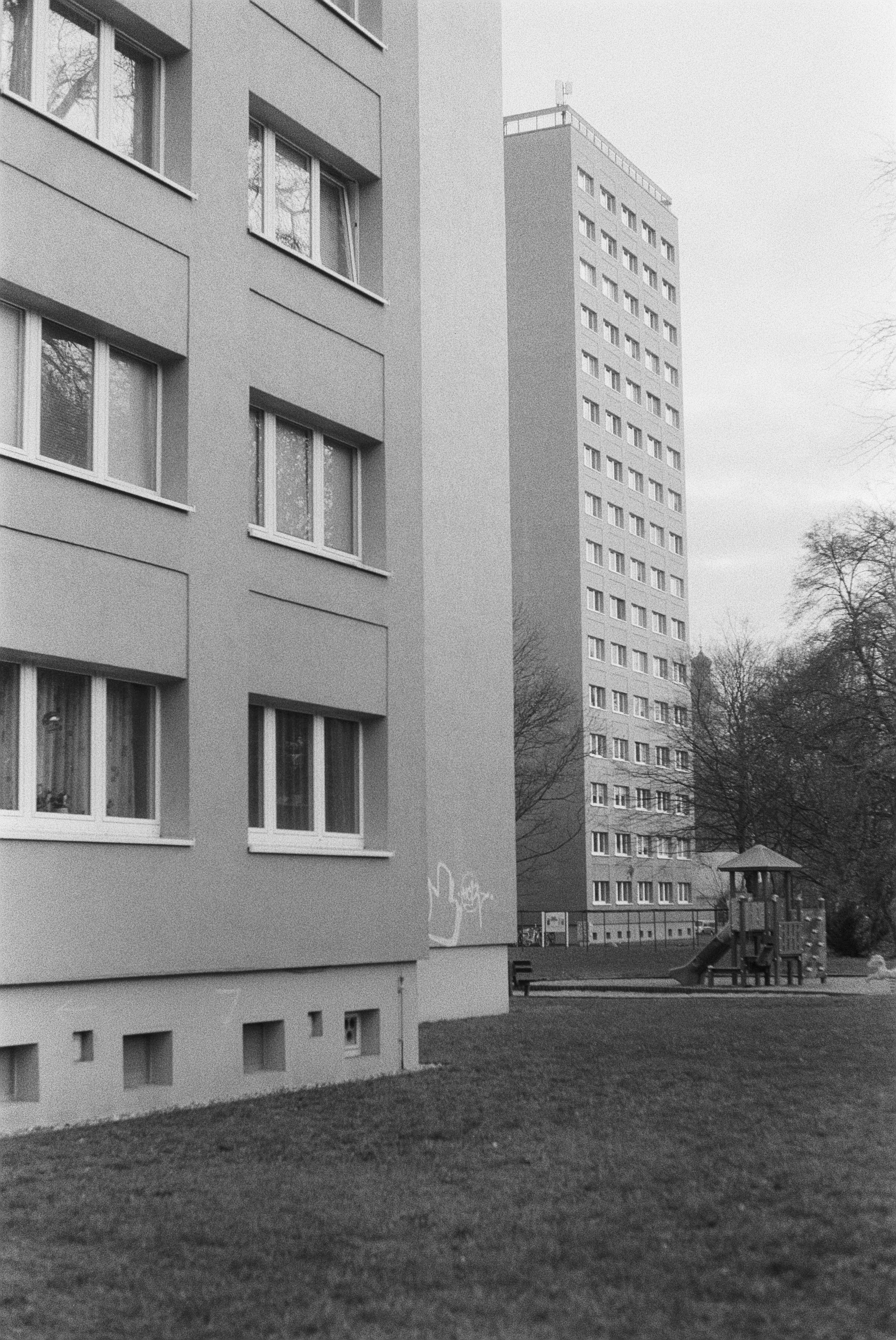 Ein mehrstöckiges Wohngebäude mit mehreren Fenstern, im Hintergrund ein zweites Hochhaus, umgeben von Bäumen und einem Spielplatz im Vordergrund.