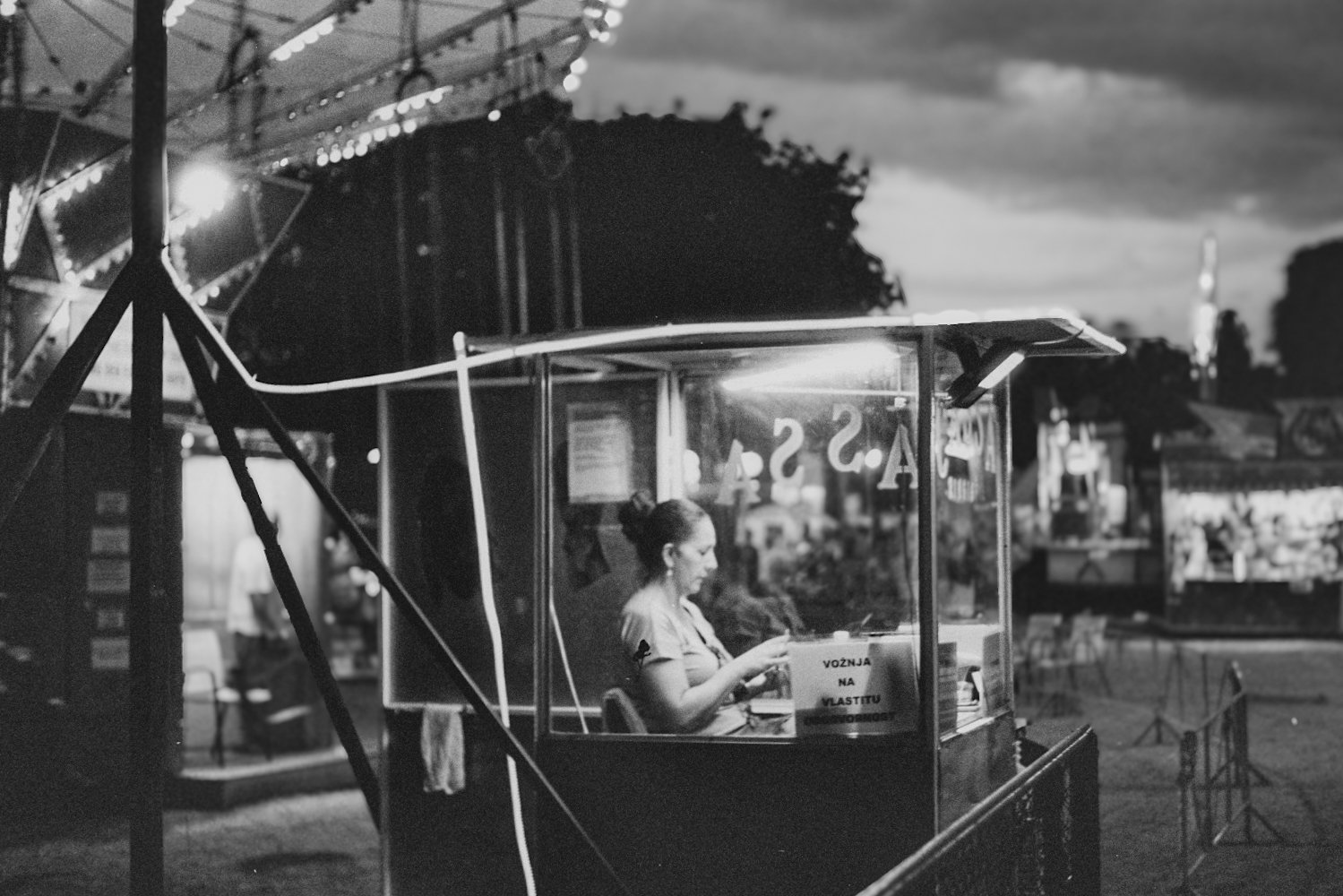 Schwarz-Weiß-Foto eines Mädchens, das in einem Fahrgeschäft vor der Dämmerung auf einem Jahrmarkt sitzt, das an einem Wagen im Inneren arbeitet.