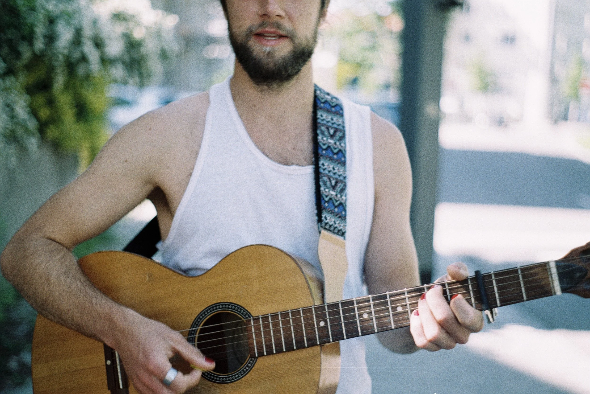 Mann spielt Gitarre im Freien, trägt ein weißes Tanktop, hat dunkles Haar und Bart, umgeben von grüner Natur mit Bäumen im Hintergrund.