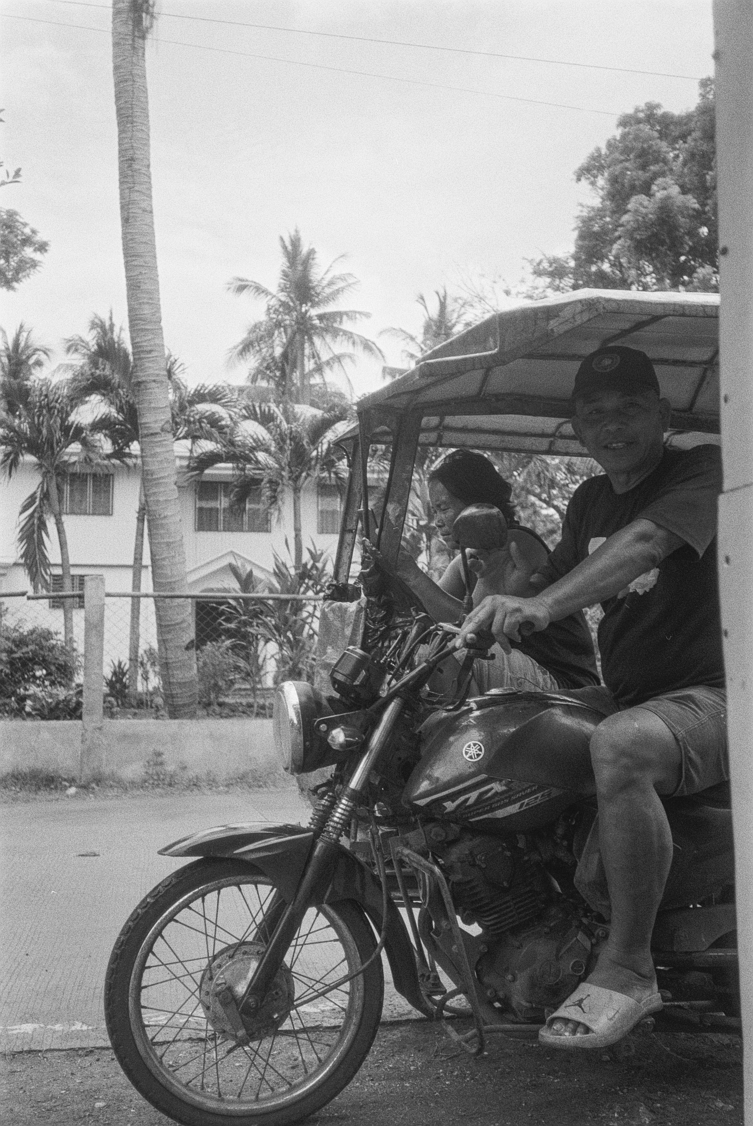 Ein Mann fährt auf einem Motorrad, während eine Frau hinter ihm sitzt und den Kopf zur Seite neigt. Sie tragen beide einen Helm, die Umgebung ist von Palmen und Bäumen geprägt, Sichtbar sind Wohnhäuser im Hintergrund. Es ist eine sonnige Szene im Fre