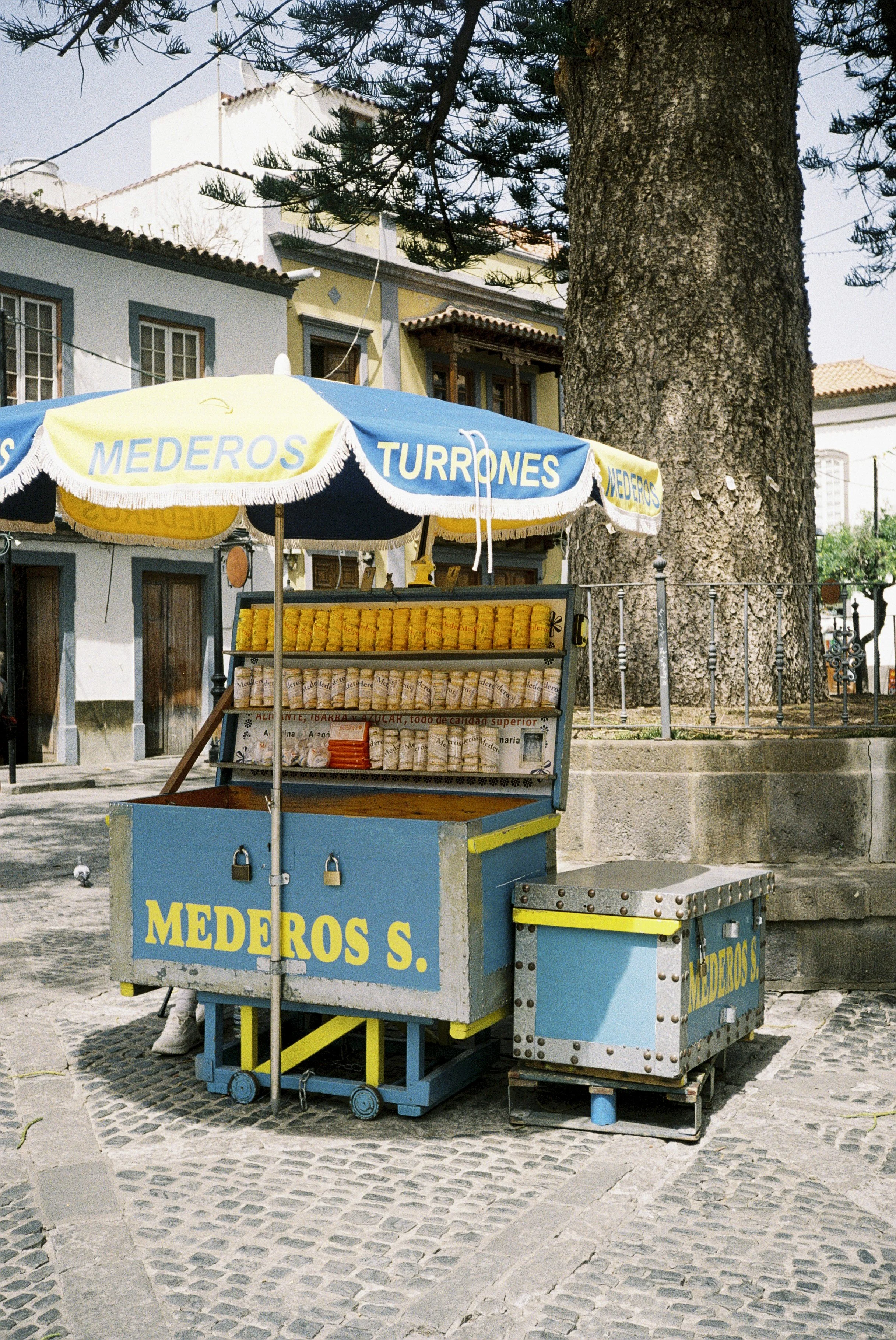 Straßenstand mit gelben und blauen Markisen, Verkaufswagen mit gelben Turrones, große Baum im Hintergrund, historische Gebäude in einer Stadt im Hintergrund, Kopf einer Taube auf dem Pflaster, typisch spanisches Stadtbild.