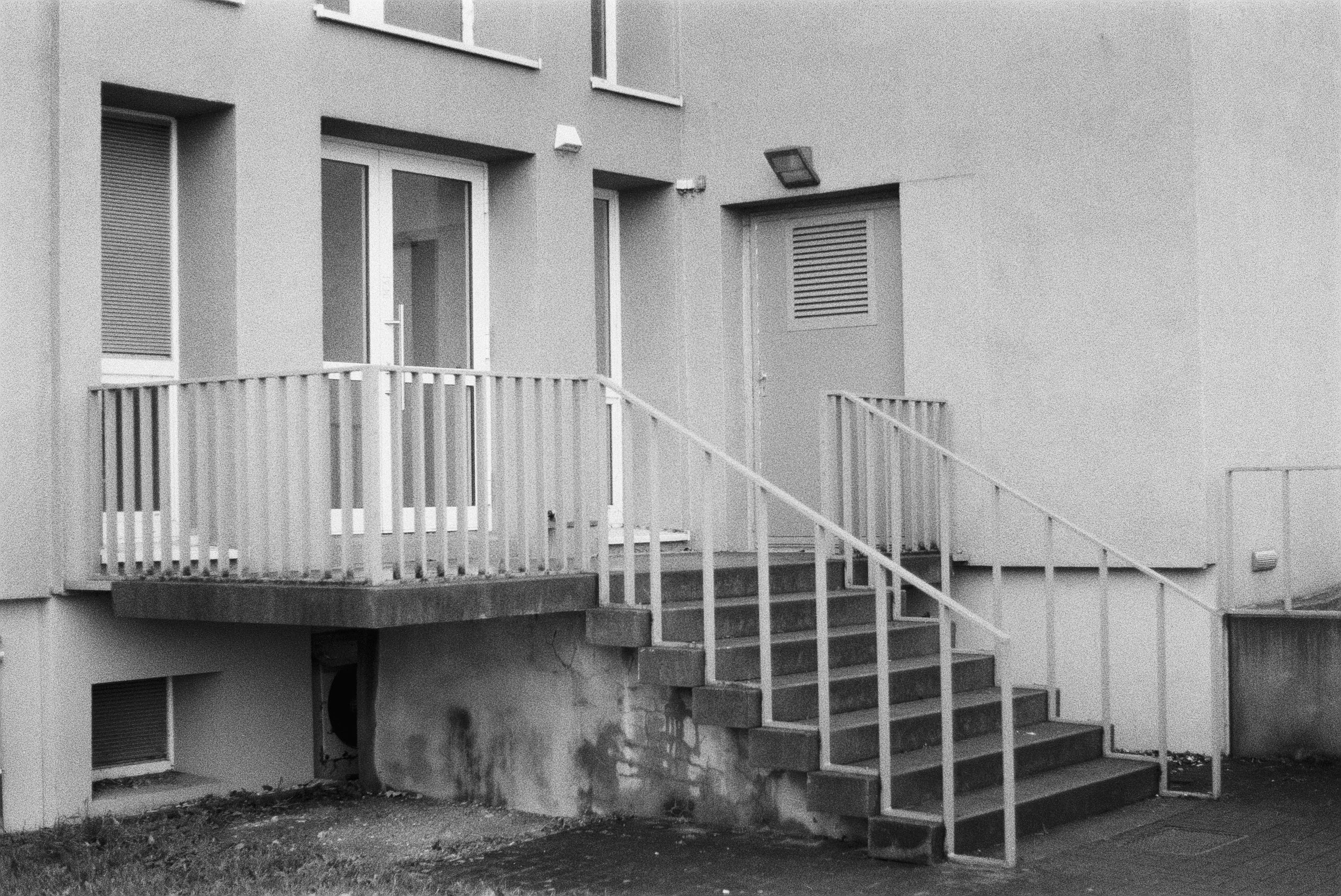 Schwarz-weiß-Foto eines Mehrfamilienhauses mit Balkon, Treppe und stehendem Baum im Vordergrund.