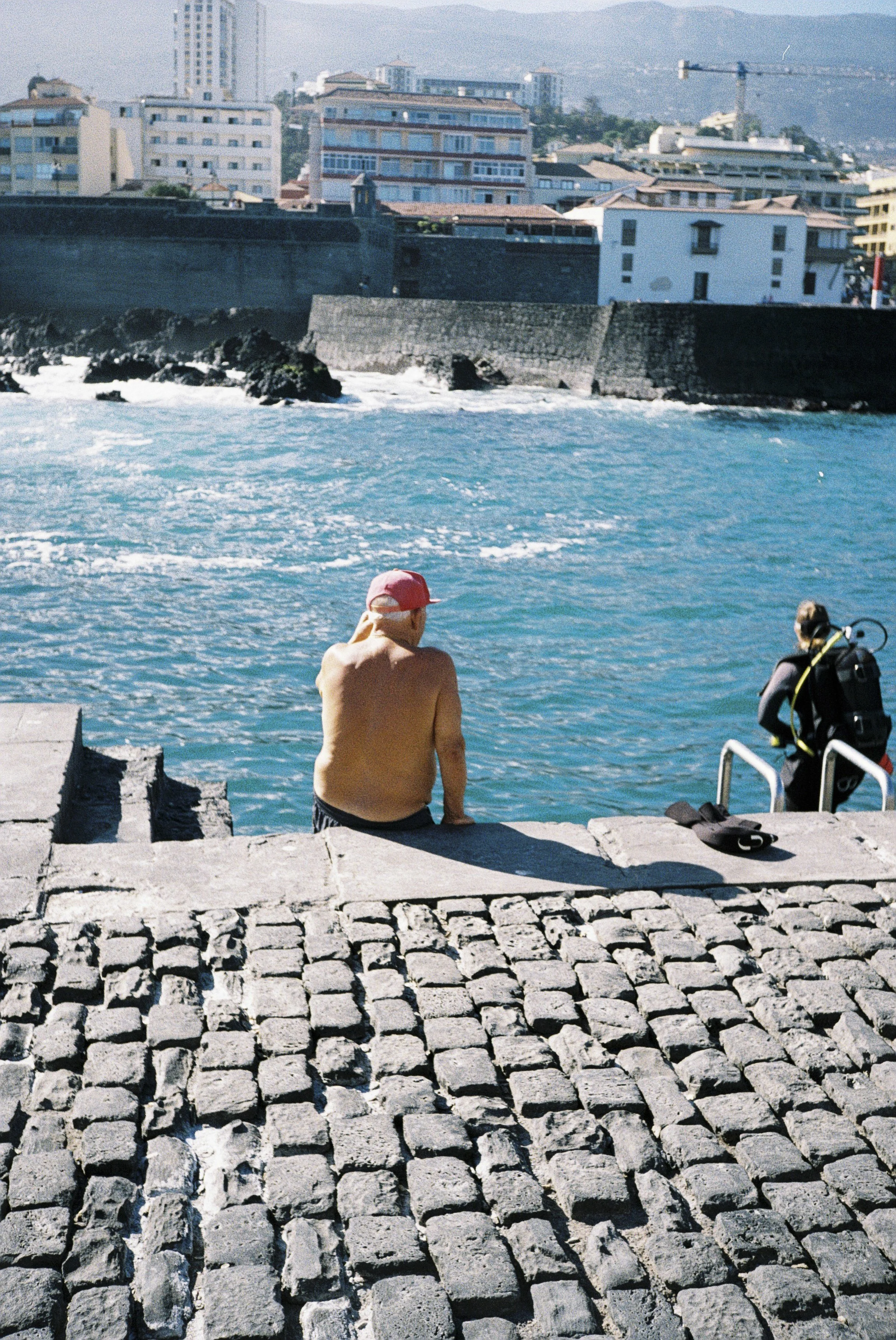 Ein Mann sitzt am Hafen, Blick auf das Wasser, mit Hochhäusern im Hintergrund.