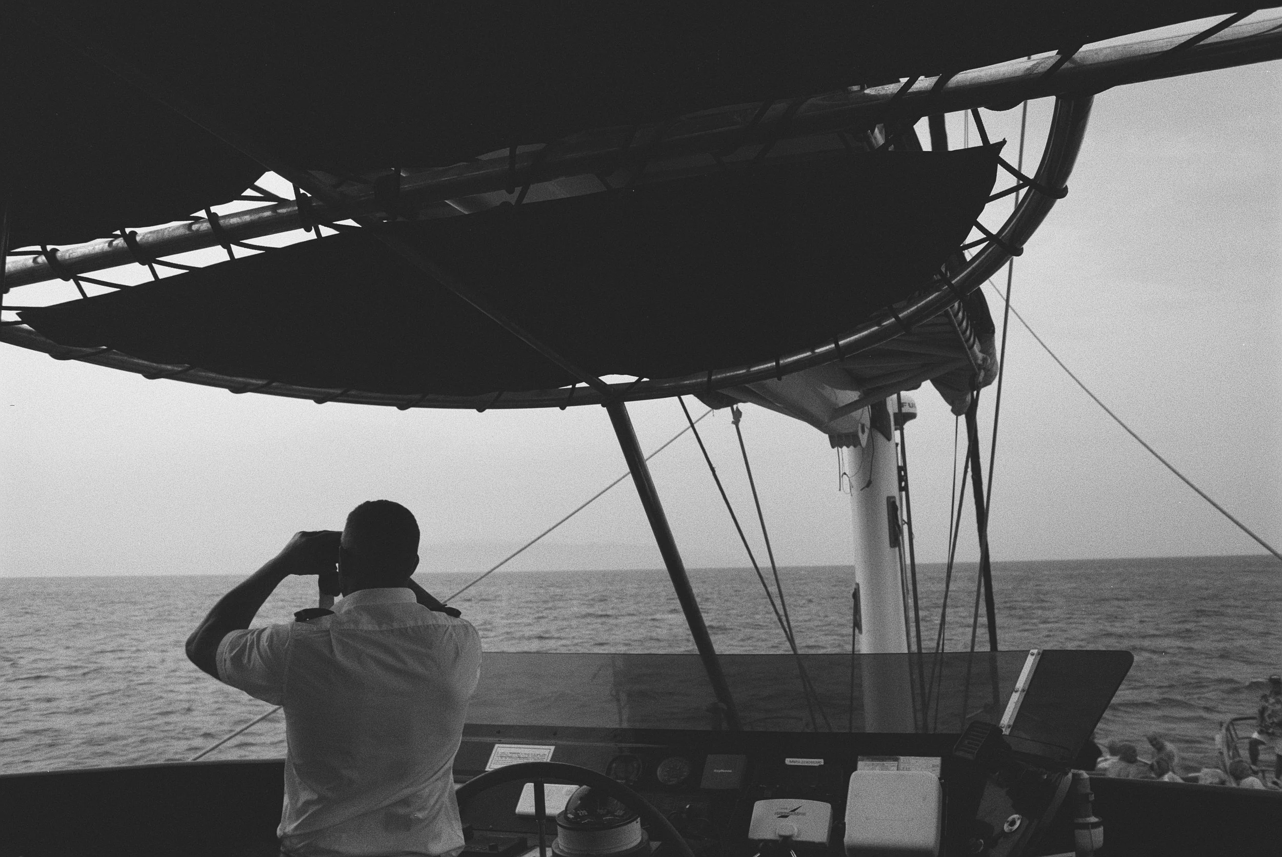Blick aus dem Steuerhaus eines Bootes auf das Meer, mit einer Person, die fotografiert, im Vordergrund. Das Boot hat ein Sonnensegel, das teilweise sichtbar ist, und es dominiert eine Schwarz-Weiß-Darstellung.