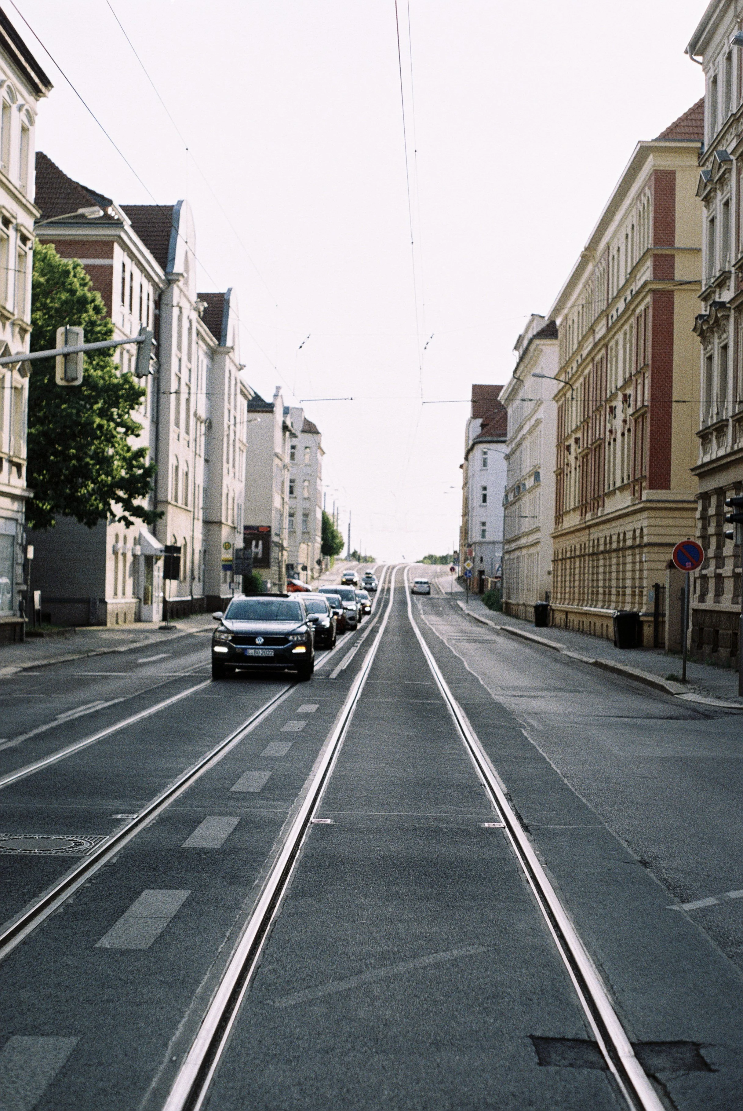 Straßenbahnlinie fährt durch eine bewohnte Straße in einer Stadt, die auf beiden Seiten von mehrstöckigen Häusern gesäumt ist, mit Autos im Vordergrund und einer etwas ansteigenden Straße im Hintergrund.