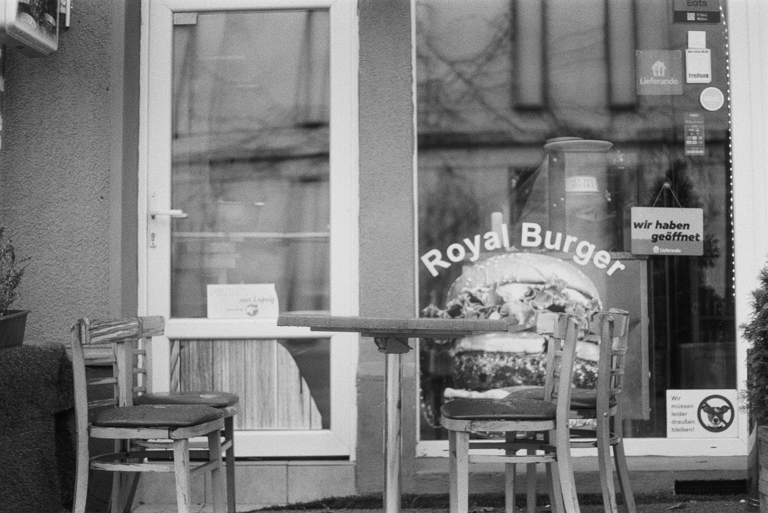 Ein Restaurant mit einem Fenster, das ein rotes Burger-Schild zeigt. Vor dem Fenster stehen Holztische und Stühle. An der Tür hängt ein Schild mit der Aufschrift 'wir haben geöffnet'.