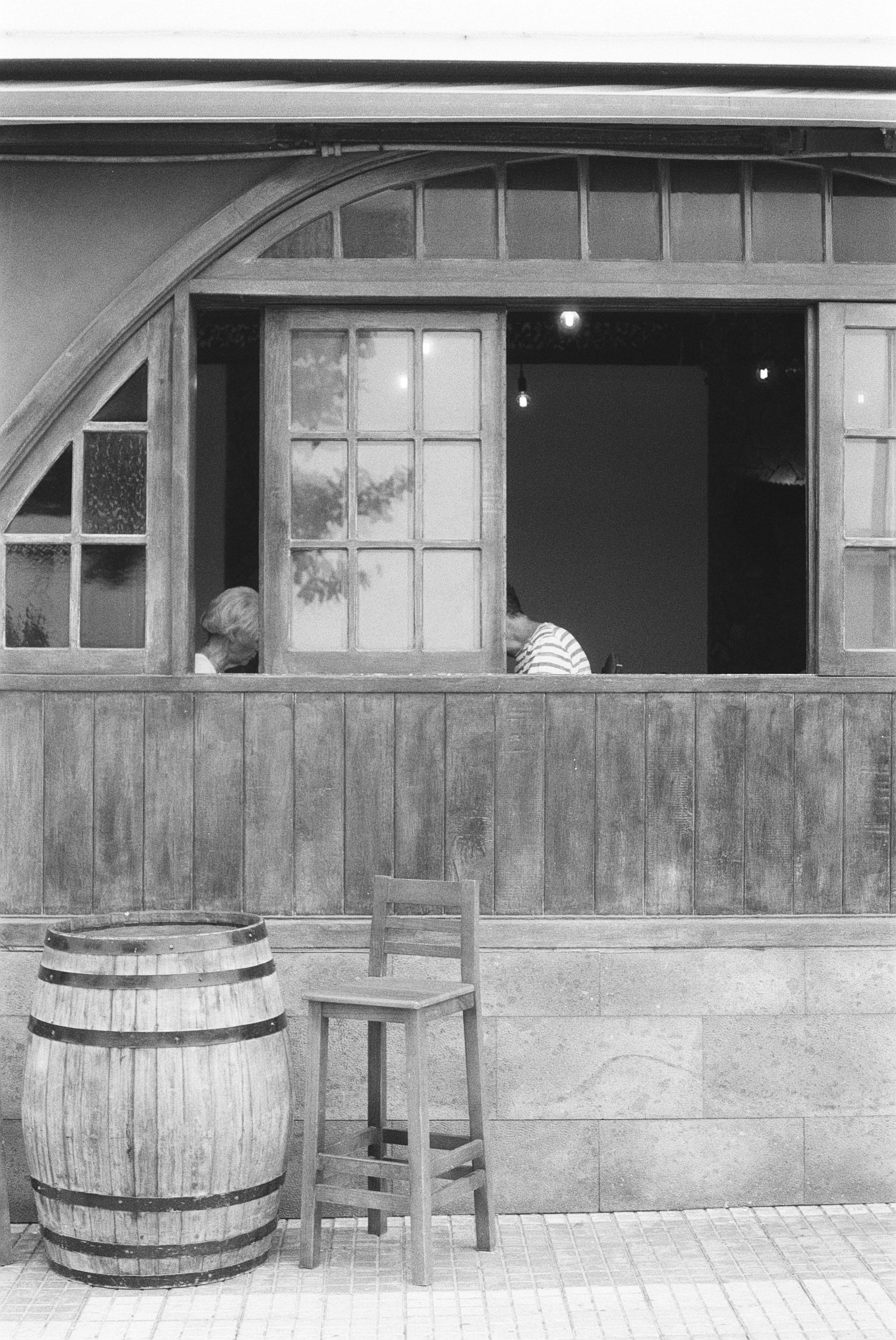 Ein Holzfass, ein Stuhl und ein Barhocker vor einer Holzwand mit einer Fensteröffnung, im Hintergrund sind zwei Menschen zu sehen.