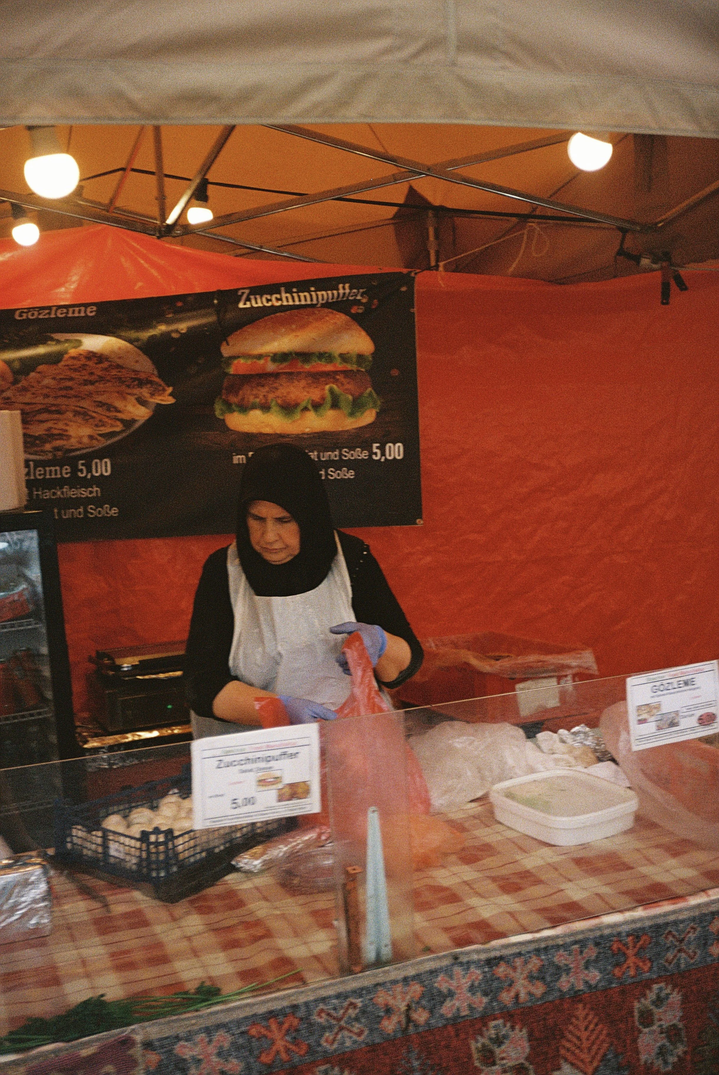 Eine Frau steht in einem warm beleuchteten Verkaufsstand und bereitet Fleisch vor. Es gibt eine Theke mit verschiedenen Lebensmitteln, darunter Fleisch, und eine Menü-Tafel, die Hamburger mit Zucchinipuffer bewirbt.