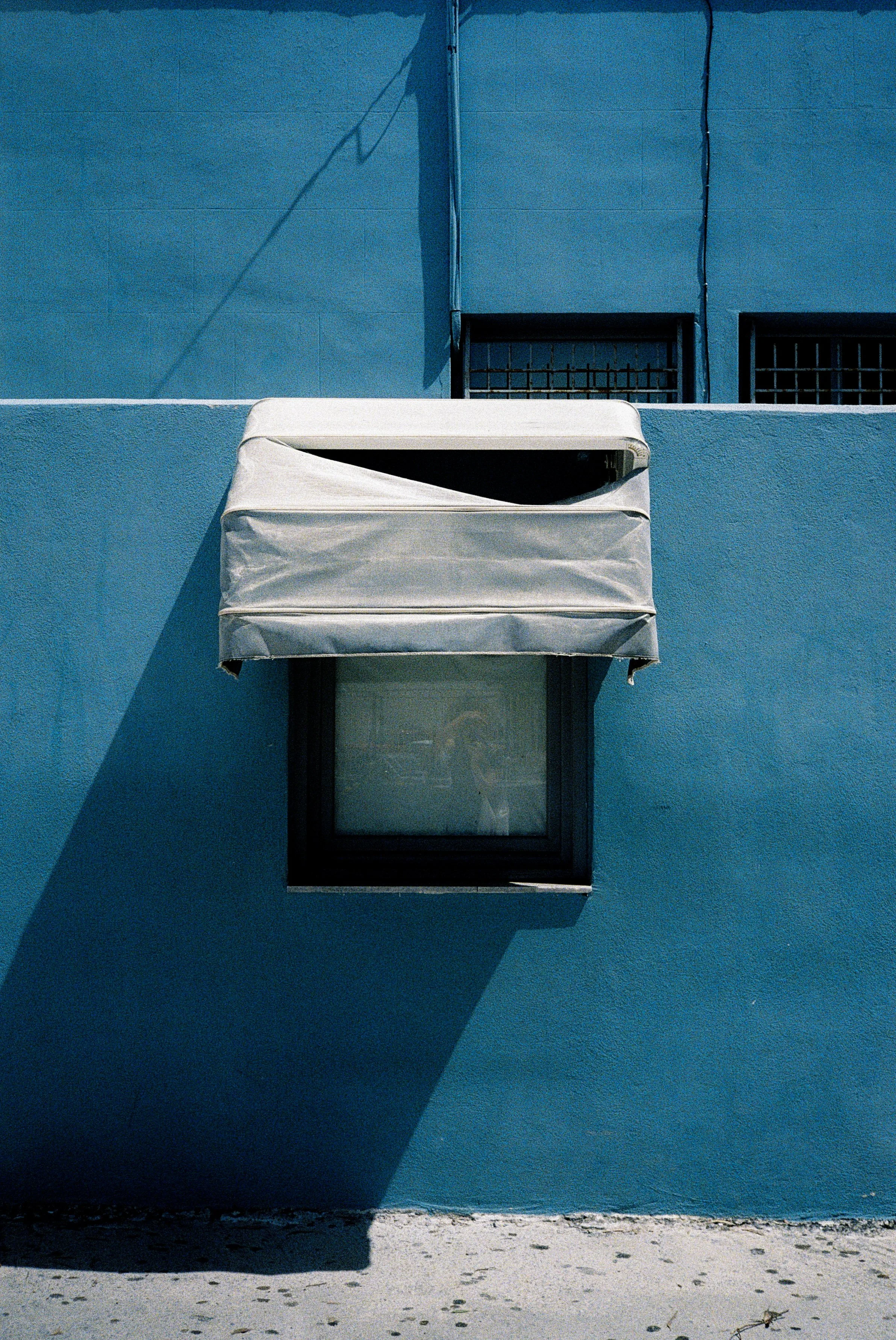 Blaue Hauswand mit zwei Fenstern, eines mit einem weißen Rolladen und das andere mit Gittern, Schatten fällt auf die Wand, kleiner Kieselsteinstrand im Vordergrund.