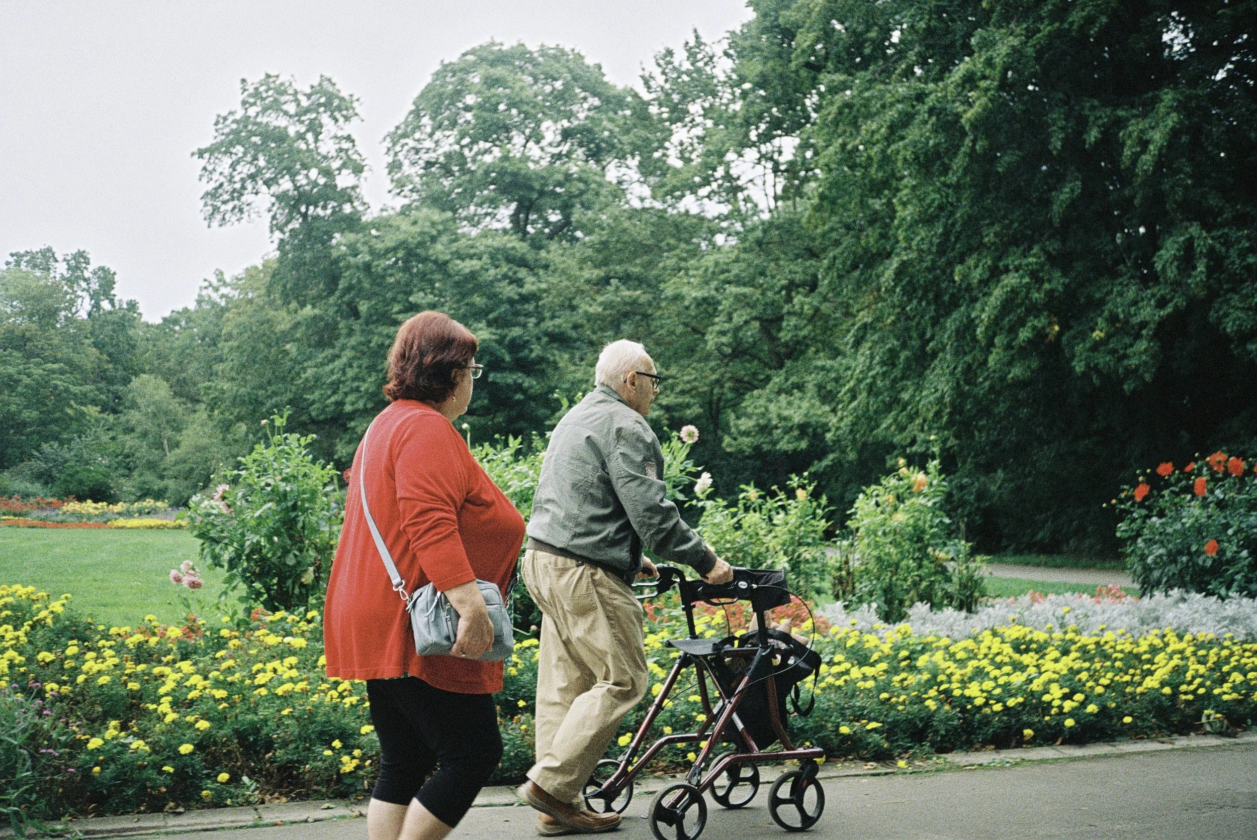 Zwei ältere Menschen, eine Frau in rotem Mantel und eine Frau mit Rollator, gehen in einem Park mit vielen Blumen und Bäumen spazieren.