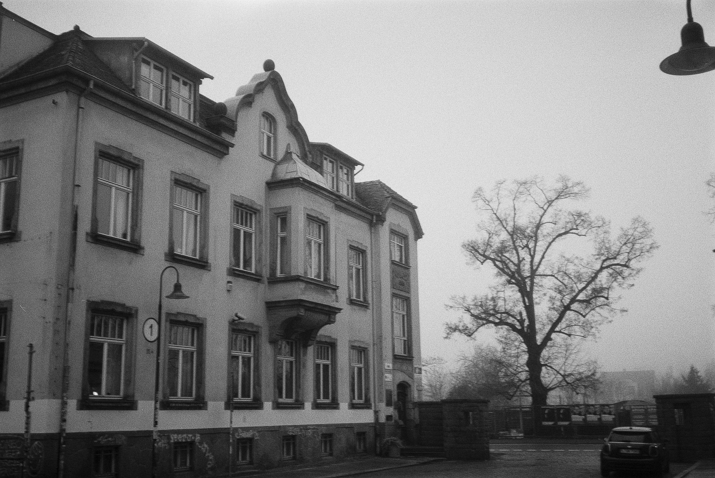 Schwarz-weiß-Aufnahme eines alten Mehrfamilienhauses mit mehreren Fenstern und Dachgauben, daneben ein großer, baum mit kaum Blättern. Im Vordergrund befindet sich ein Parkplatz mit Autos.