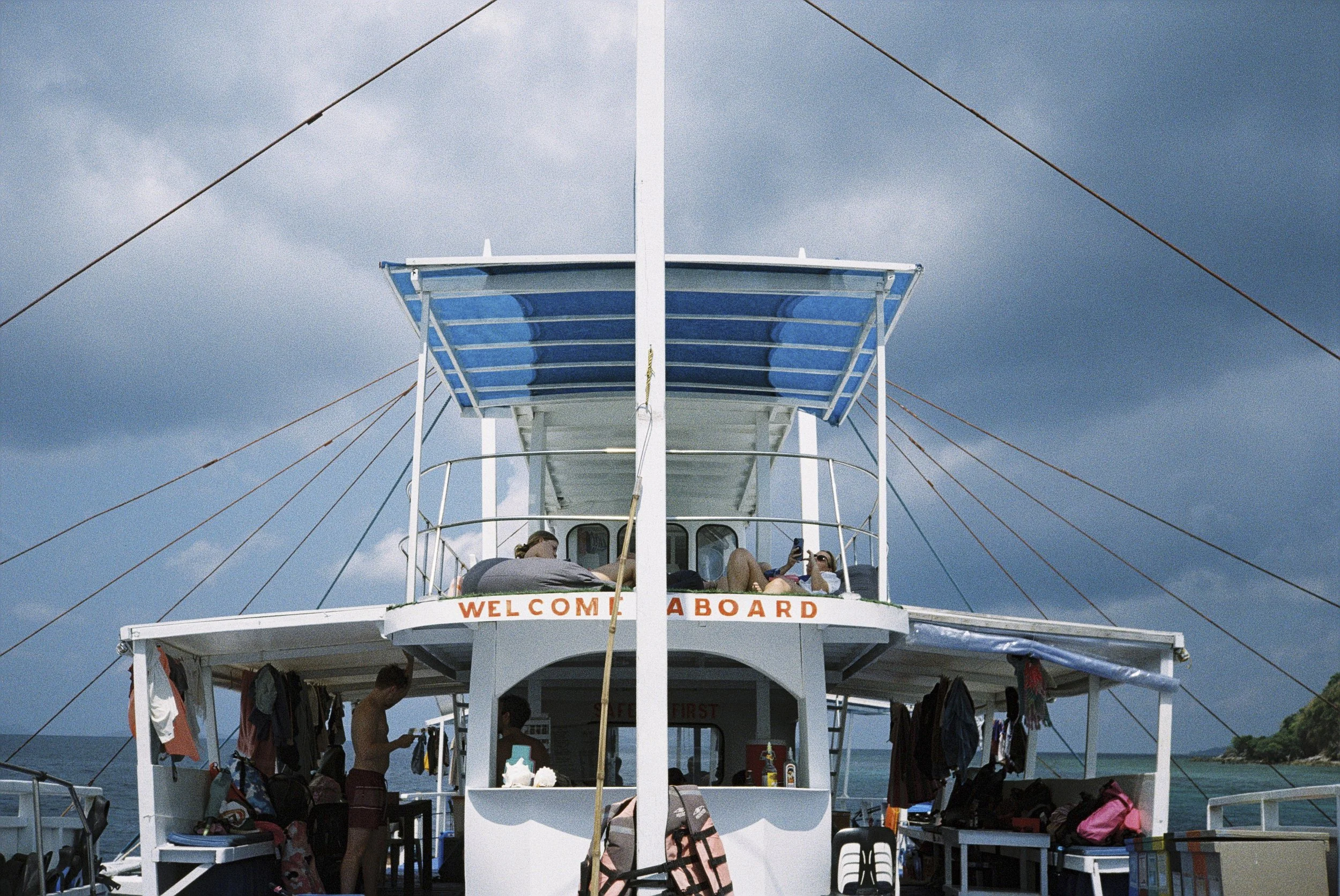 Menschen entspannen sich auf einem weißen Boot mit einem oberen Deck und stützen sich auf Geländer, während sie ins Meer blicken, bei bewölktem Himmel.