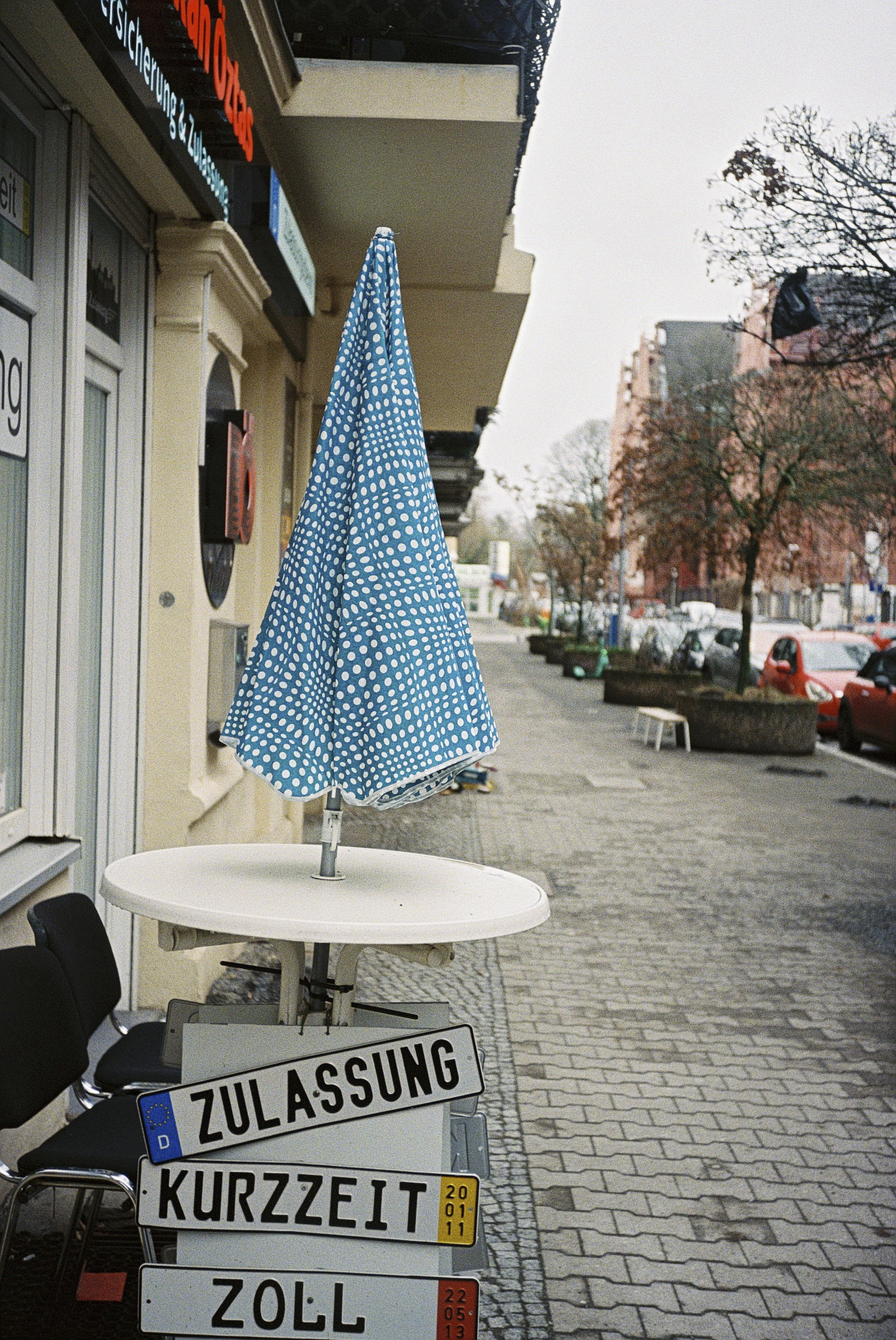 Auf der linken Seite stehen mehrere Verkehrsschilder mit deutschen Beschriftungen, darunter 'Zulassung', 'Kurzzeit', 'Zoll'. Im Vordergrund ist ein weißer Tisch mit einem blauen, gepunkteten Sonnenschirm zu sehen, der auf einer öffentlichen Straße st