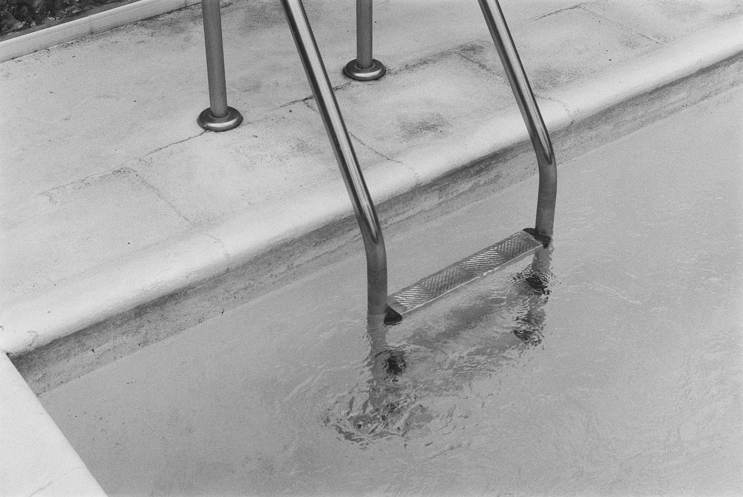 Ein Treppengeländer am Rand eines Schwimmbeckens, das Wasser spiegelt sich im Becken. Das Bild ist in Schwarz-Weiß.