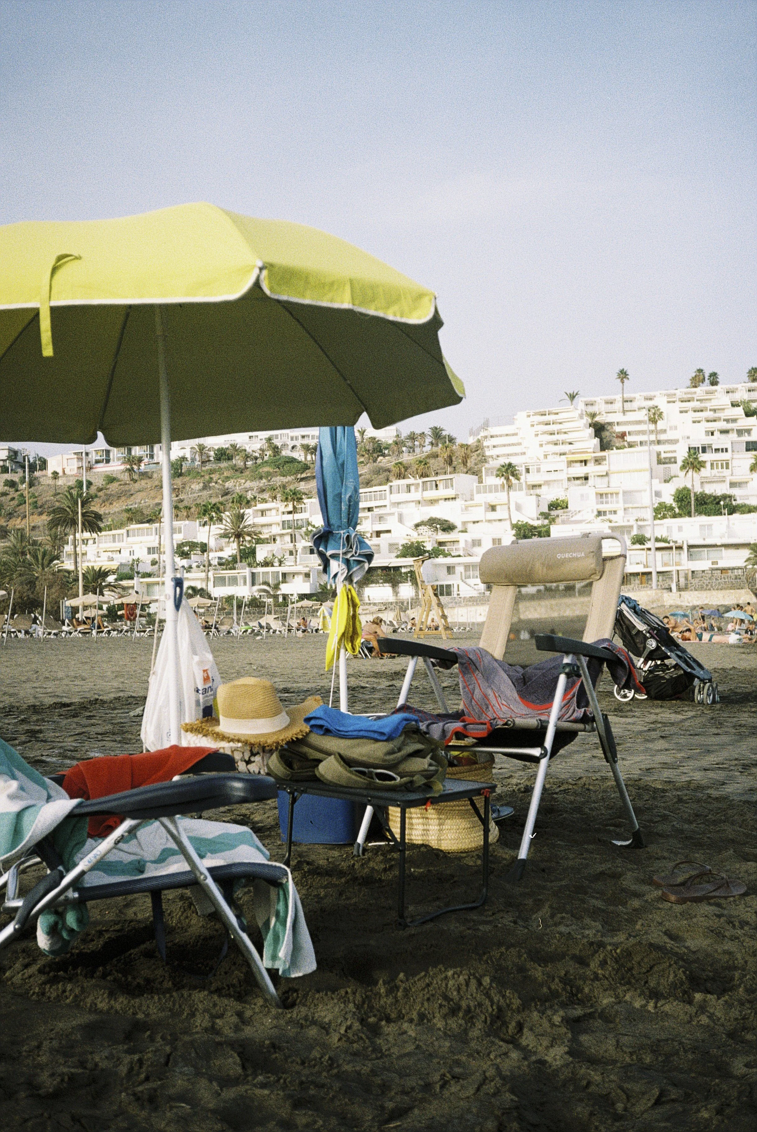 Strand mit grüner Sonnenbank, Sonnenschirm, Liegen, Strandtaschen und Hüten, mit weißen Gebäuden und Palmen im Hintergrund bei sonnigem Wetter.