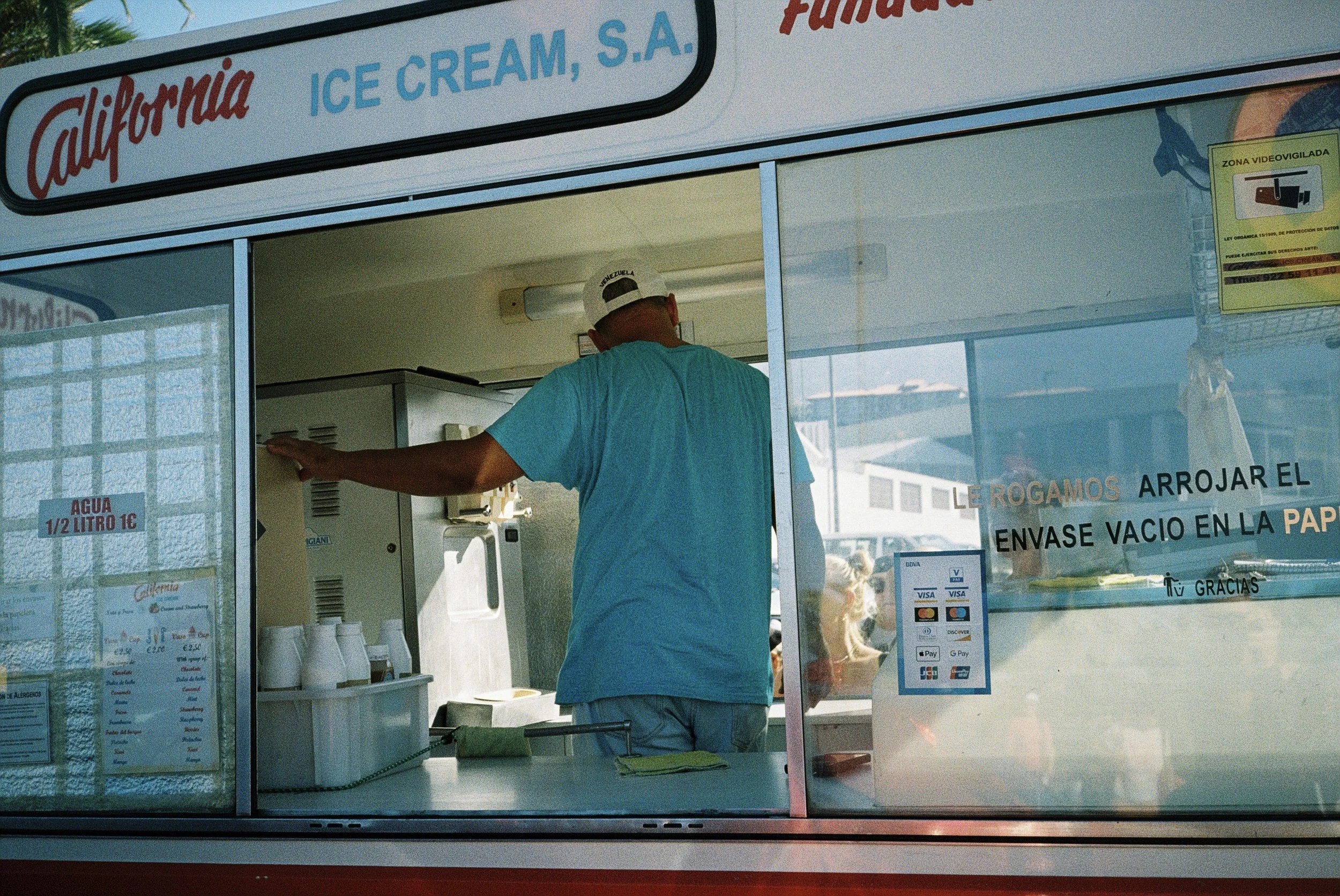 Ein Verkäufer in einem Imbisswagen backt oder bereitet Essen vor, während Kunden draußen warten, zu sehen durch das Fenster. Der Imbisswagen hat ein Schild mit dem Text "California Ice Cream" und weiteren Informationen auf spanisch.