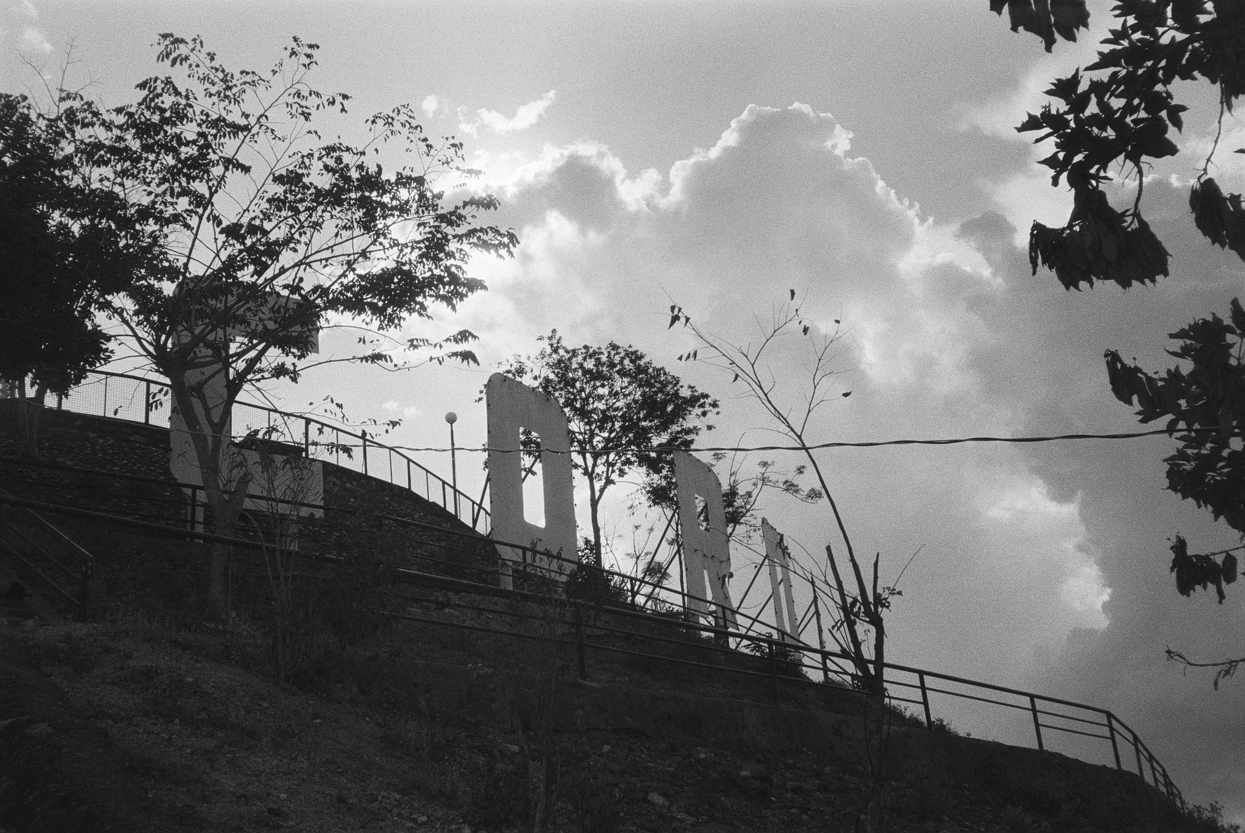 Schwarz-weiß-Foto von Bäumen auf einem Hügel mit Zaun und einer großen Buchstaben-Statue im Hintergrund, Wolken im Himmel
