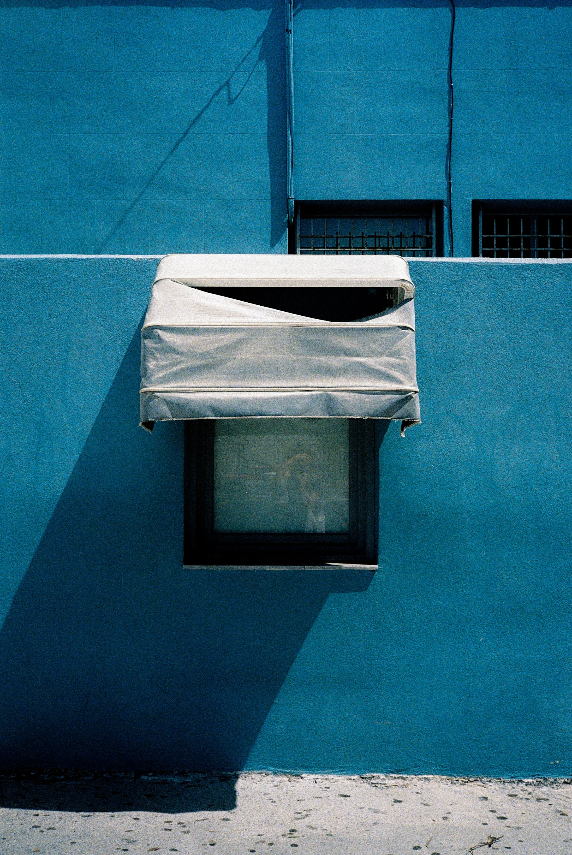 Ein blauer Gebäudeteil mit zwei Fenstern, eines mit Gitter und eines ohne. Ein Fenster hat eine weiße Gardine, die nach oben weggeklappt ist. Ein Schatten wird von den Elementen auf den blauen Wänden geworfen. Der Boden ist sandig oder staubig.